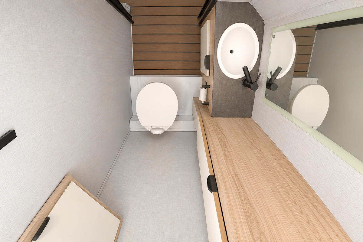 Van interior with toilet, sink, large mirror, wooden countertop, cabinet, and white walls.