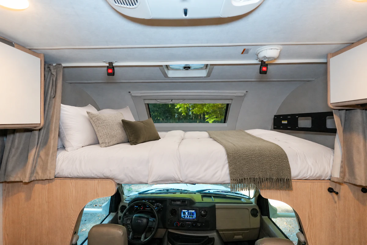 Over-cab sleeping area with double bed, pillows, blankets, overhead cabinets, small window, and view of driver’s cab.
