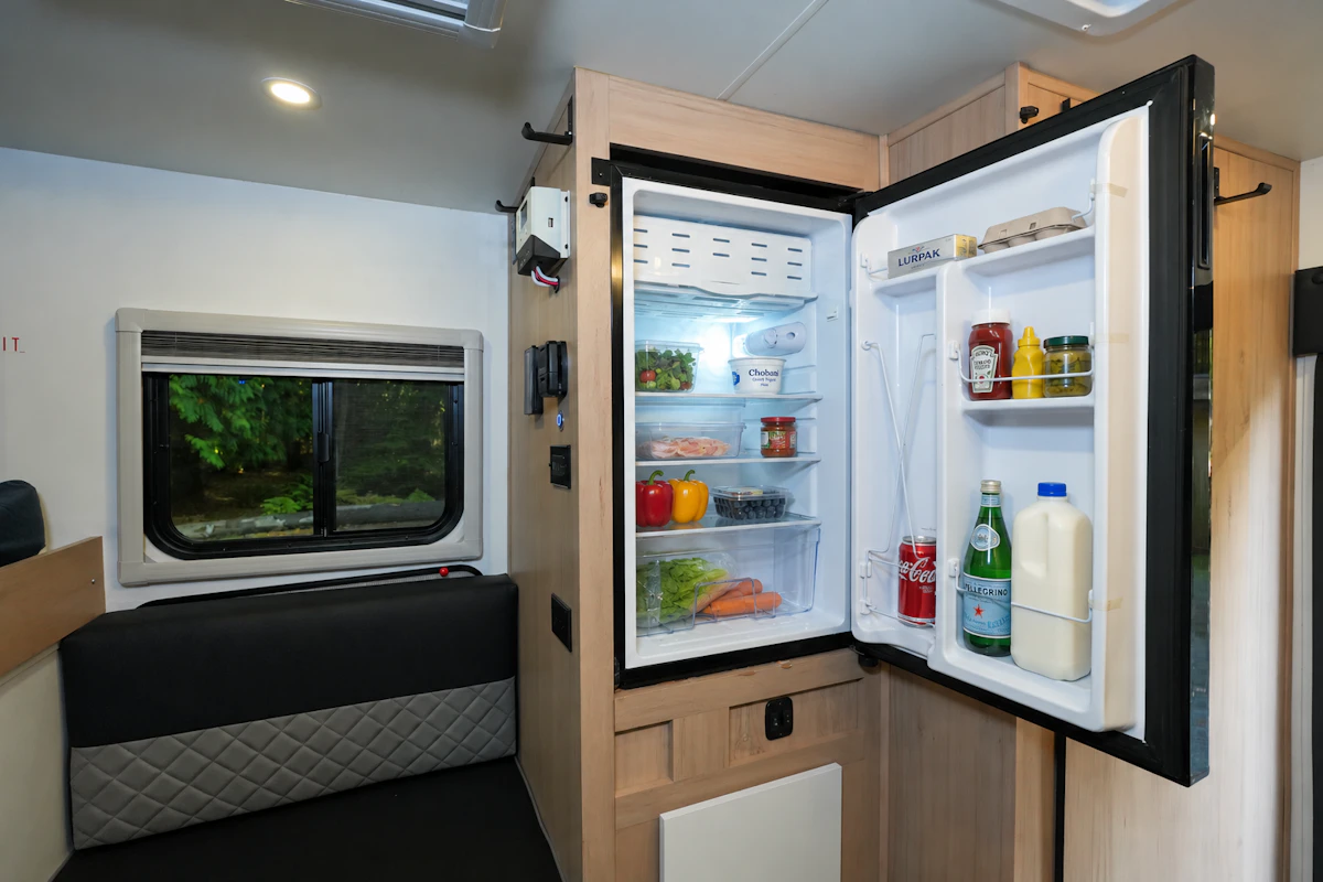Van interior with open fridge, shelves stocked with food and drinks, window, bench seat, and light wood cabinetry.