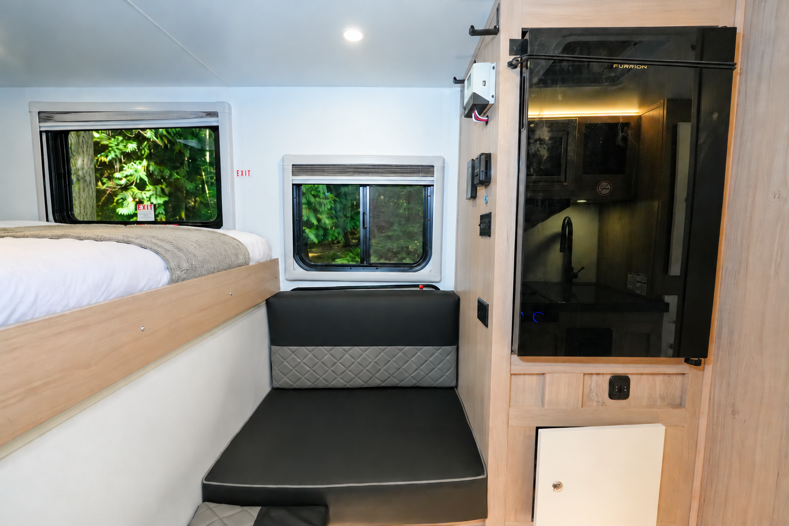 Van interior with bed above seating area, two windows, kitchen area with sink, and cabinetry visible.