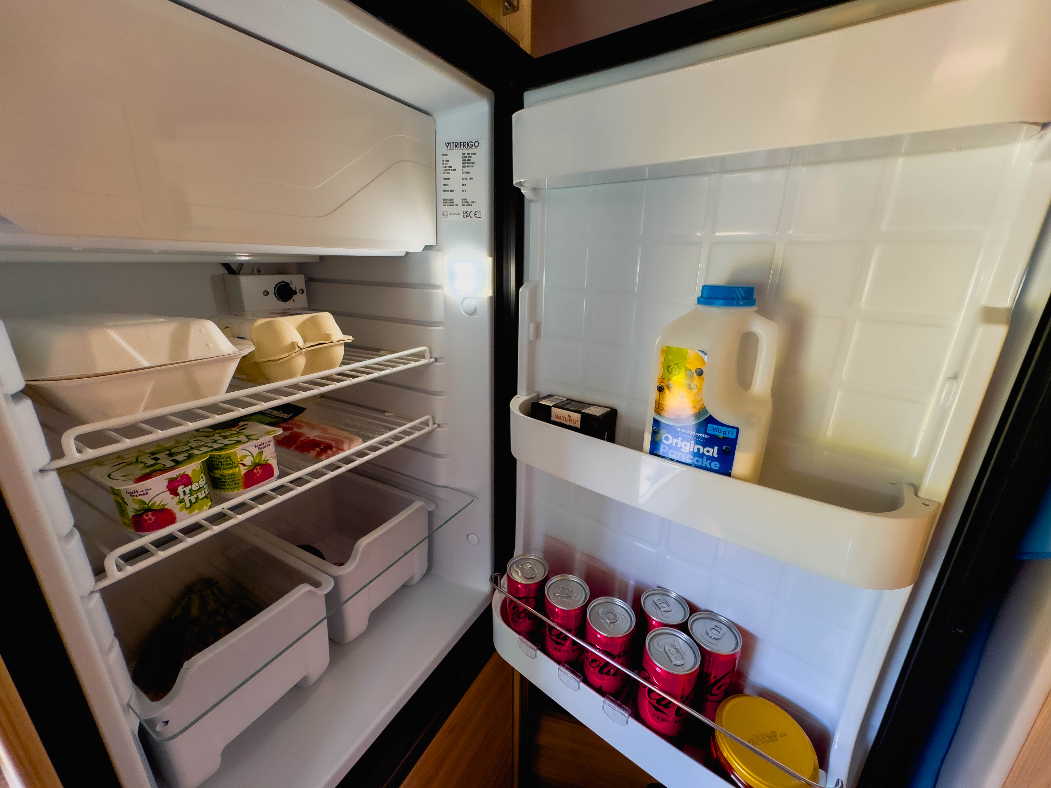 Open campervan fridge stocked with eggs, milk, canned drinks, yogurt, vegetables, and other food items on shelves.