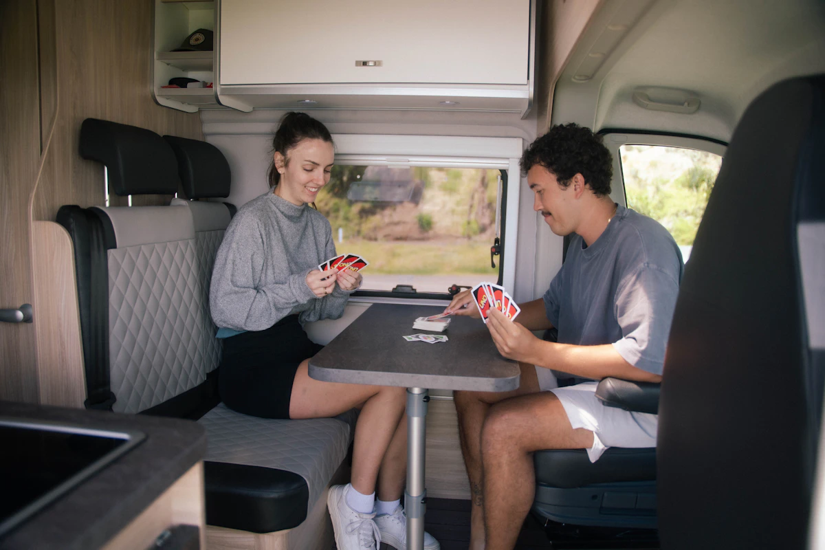 Two people sitting inside a campervan, playing a card game at the dining table during daylight.