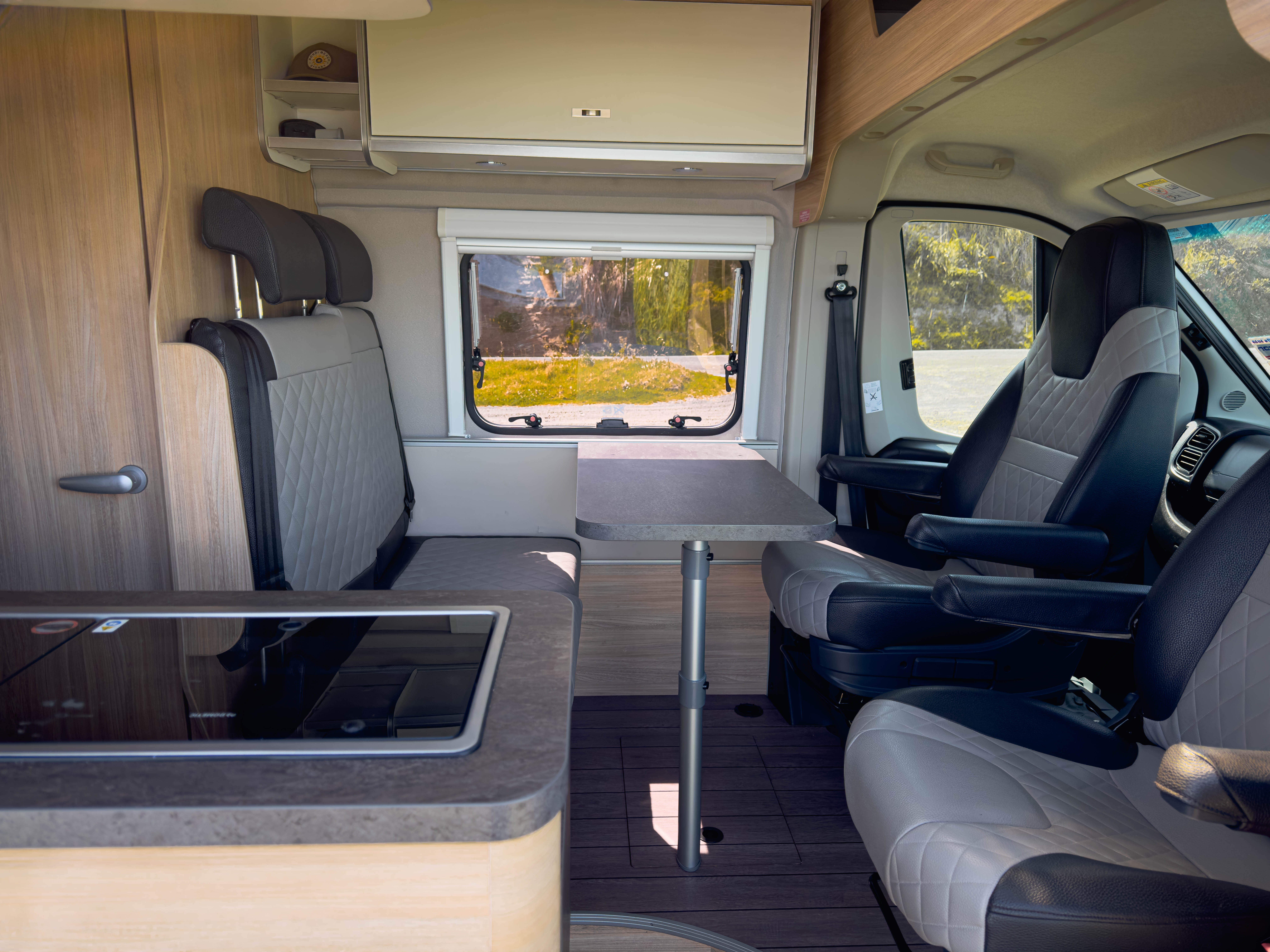 Modern campervan interior with swivel seats, dining table, kitchenette, and window view of nature in the background.