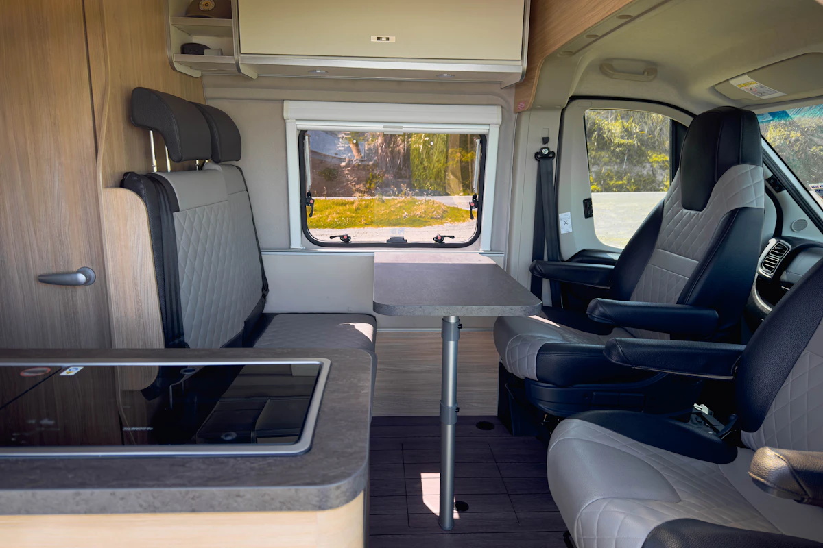 Modern campervan interior with swivel seats, dining table, kitchenette, and window view of nature in the background.