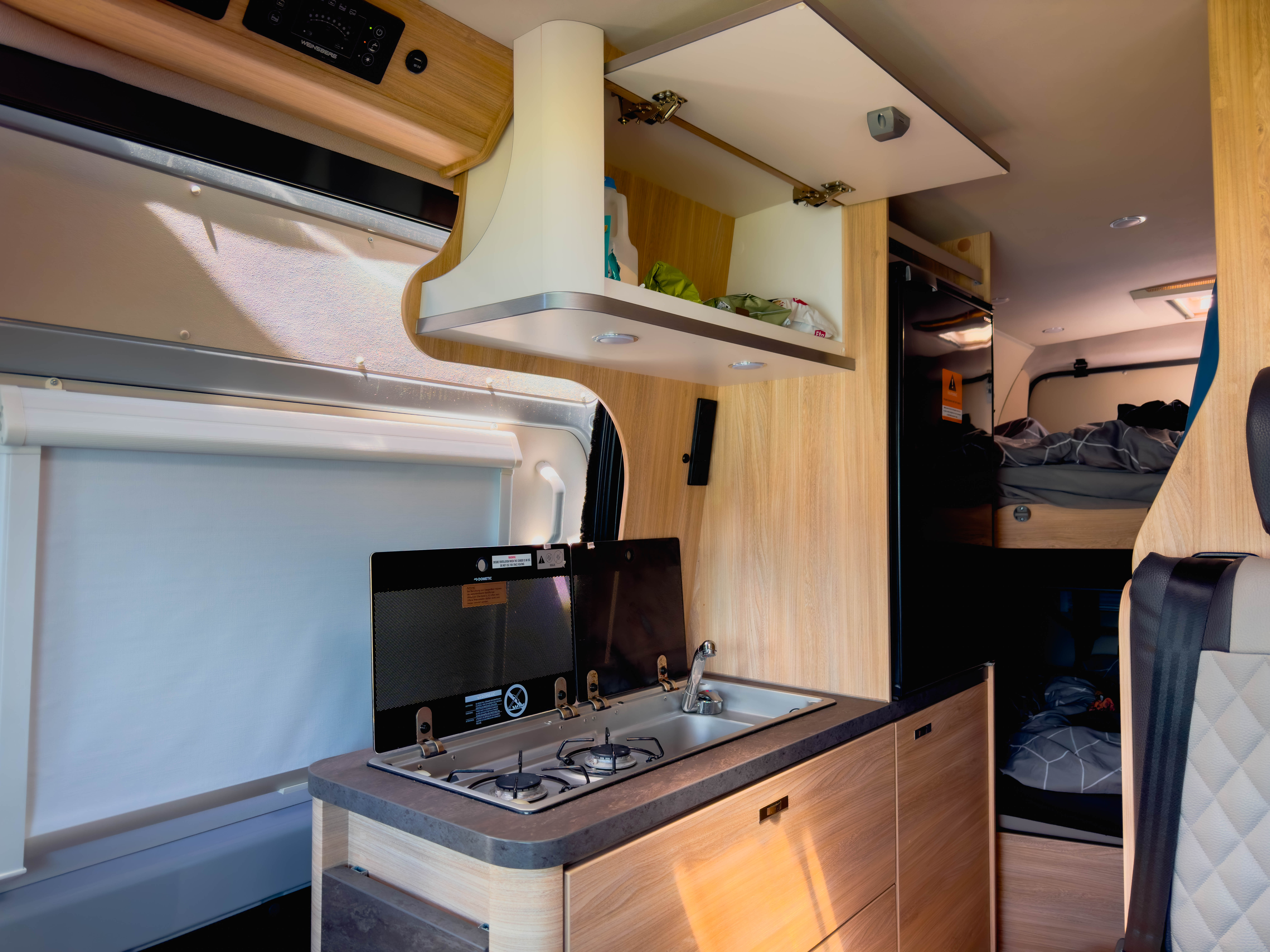 Modern campervan interior with compact kitchen, stove, sink, overhead storage, and a visible sleeping area in the background.