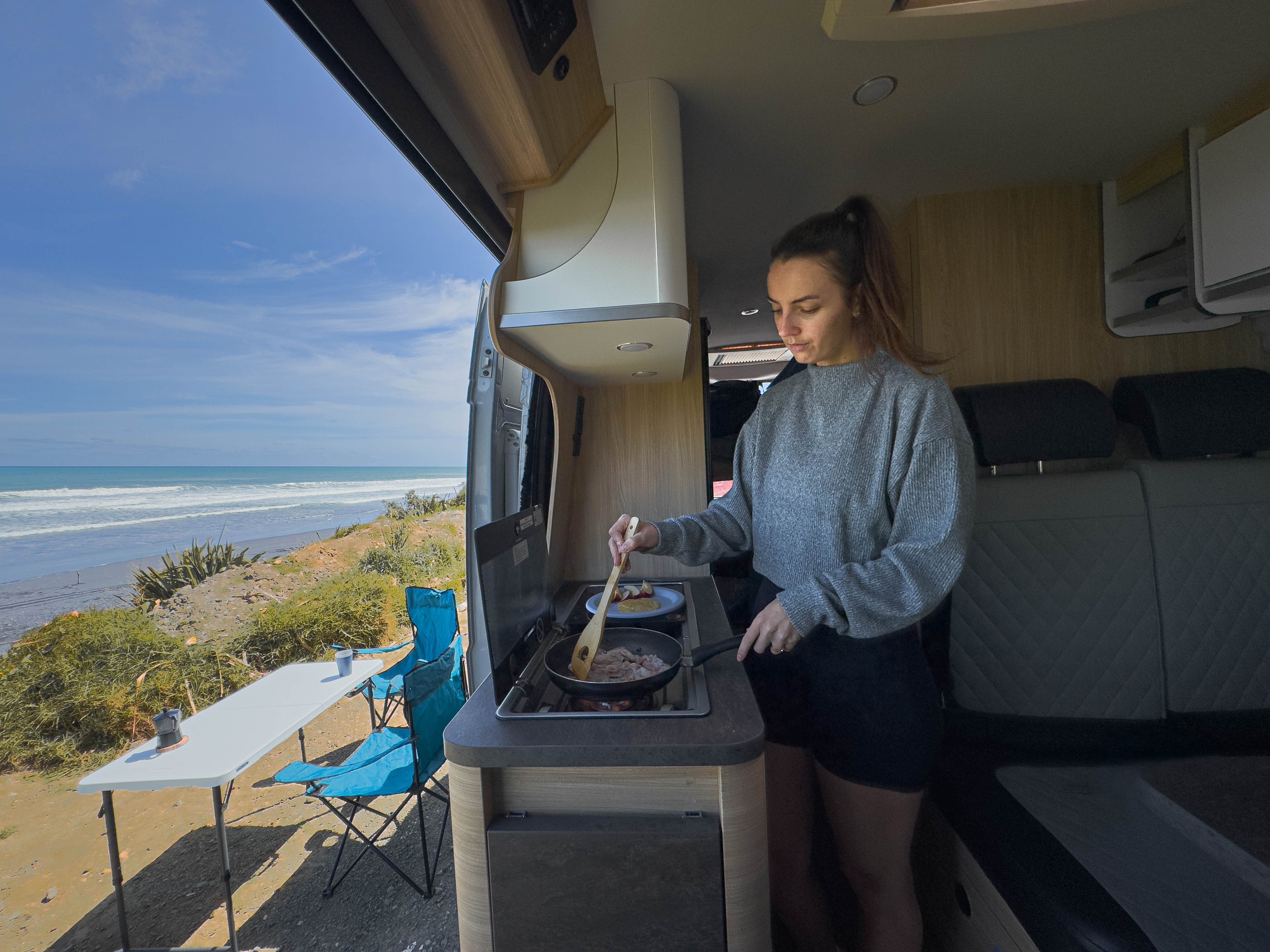 Person cooking inside a campervan parked by a beach, with outdoor table and sea view visible.