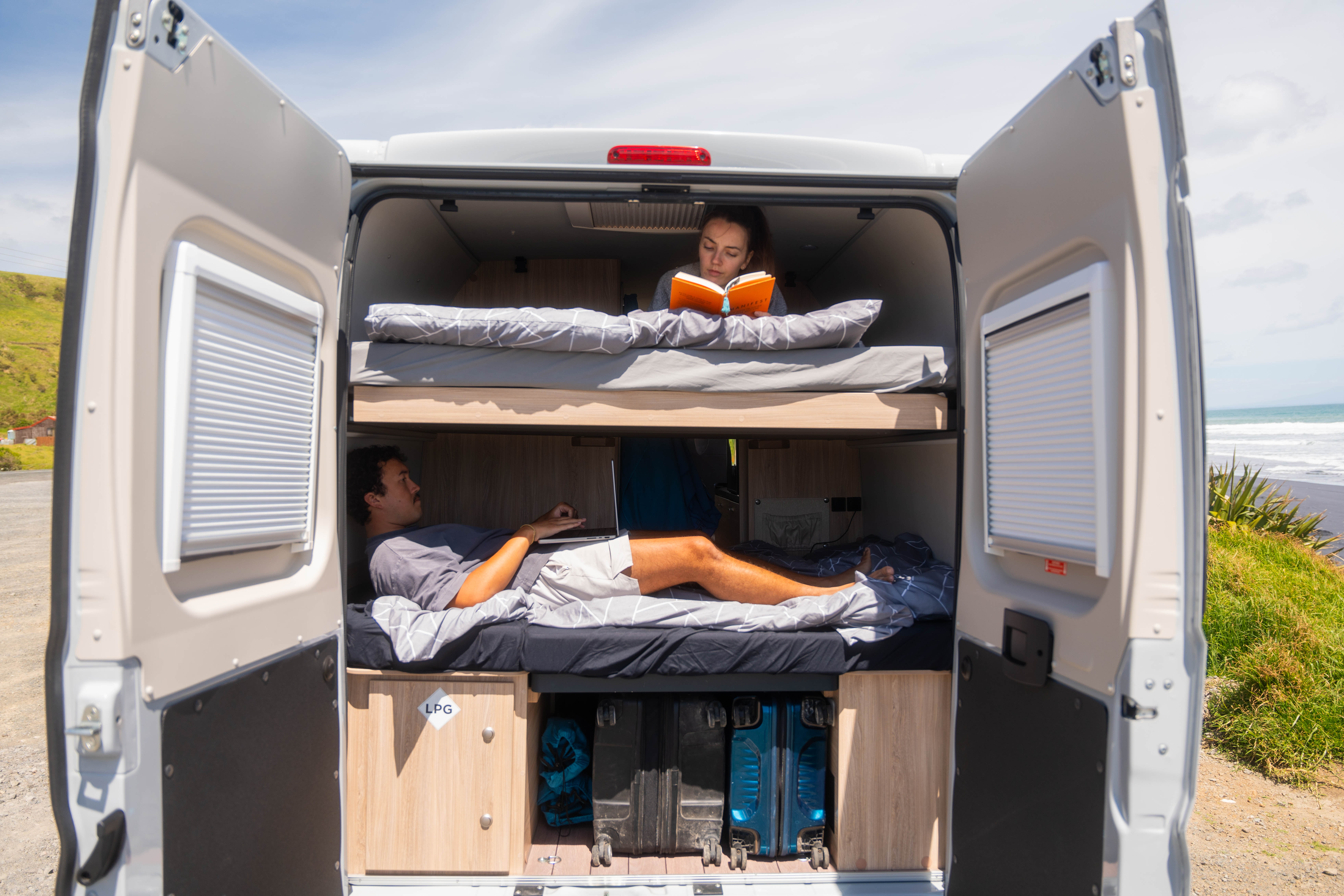 Two people relax in bunk beds inside a campervan parked near the coast, doors open to nature.