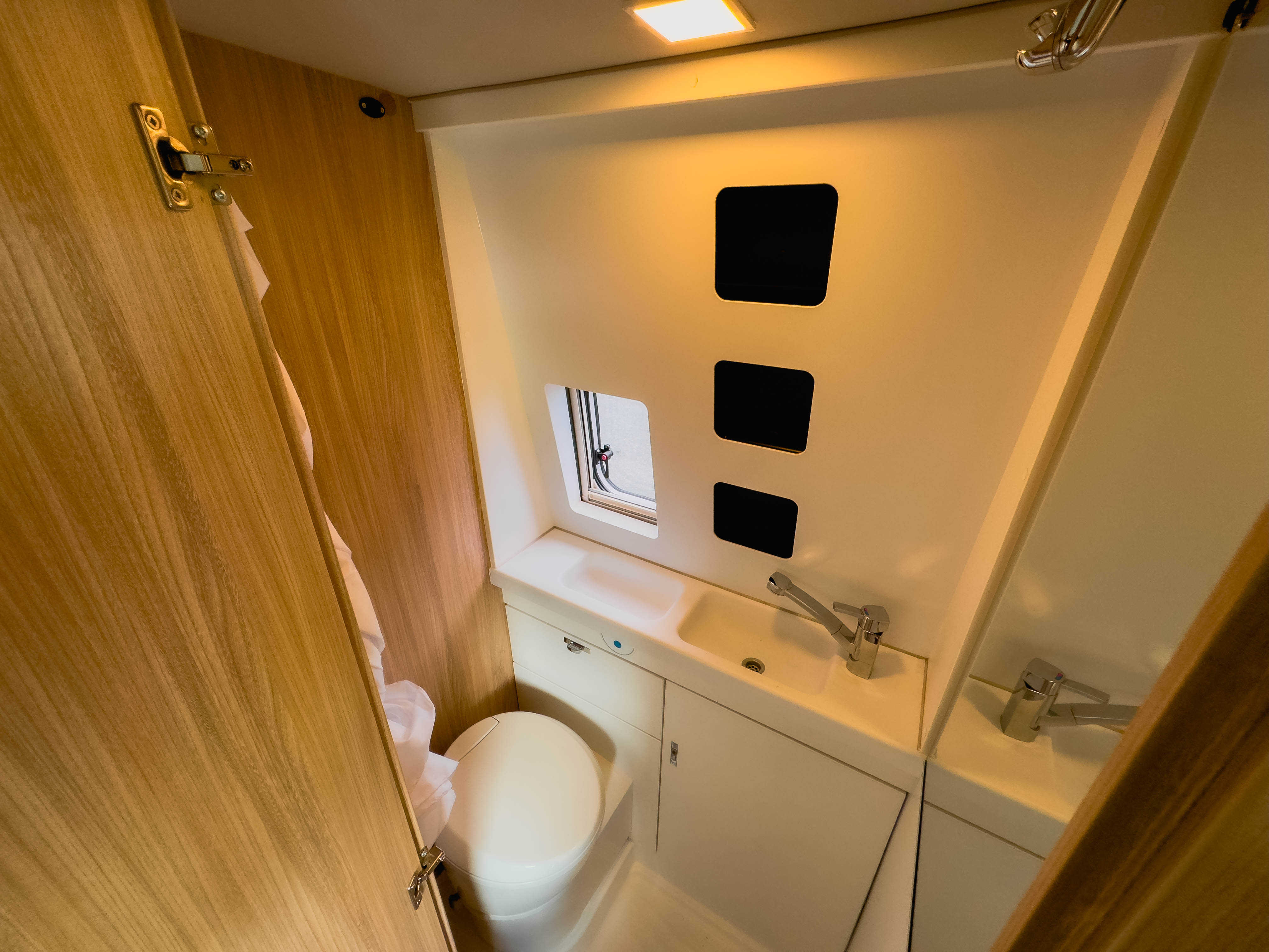 Compact campervan bathroom with toilet, sink, and small window, featuring wooden and white finishes for a modern look.