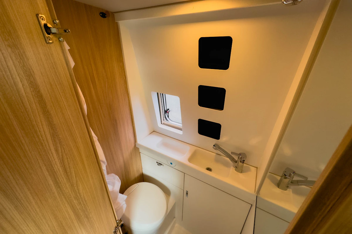Compact campervan bathroom with toilet, sink, and small window, featuring wooden and white finishes for a modern look.