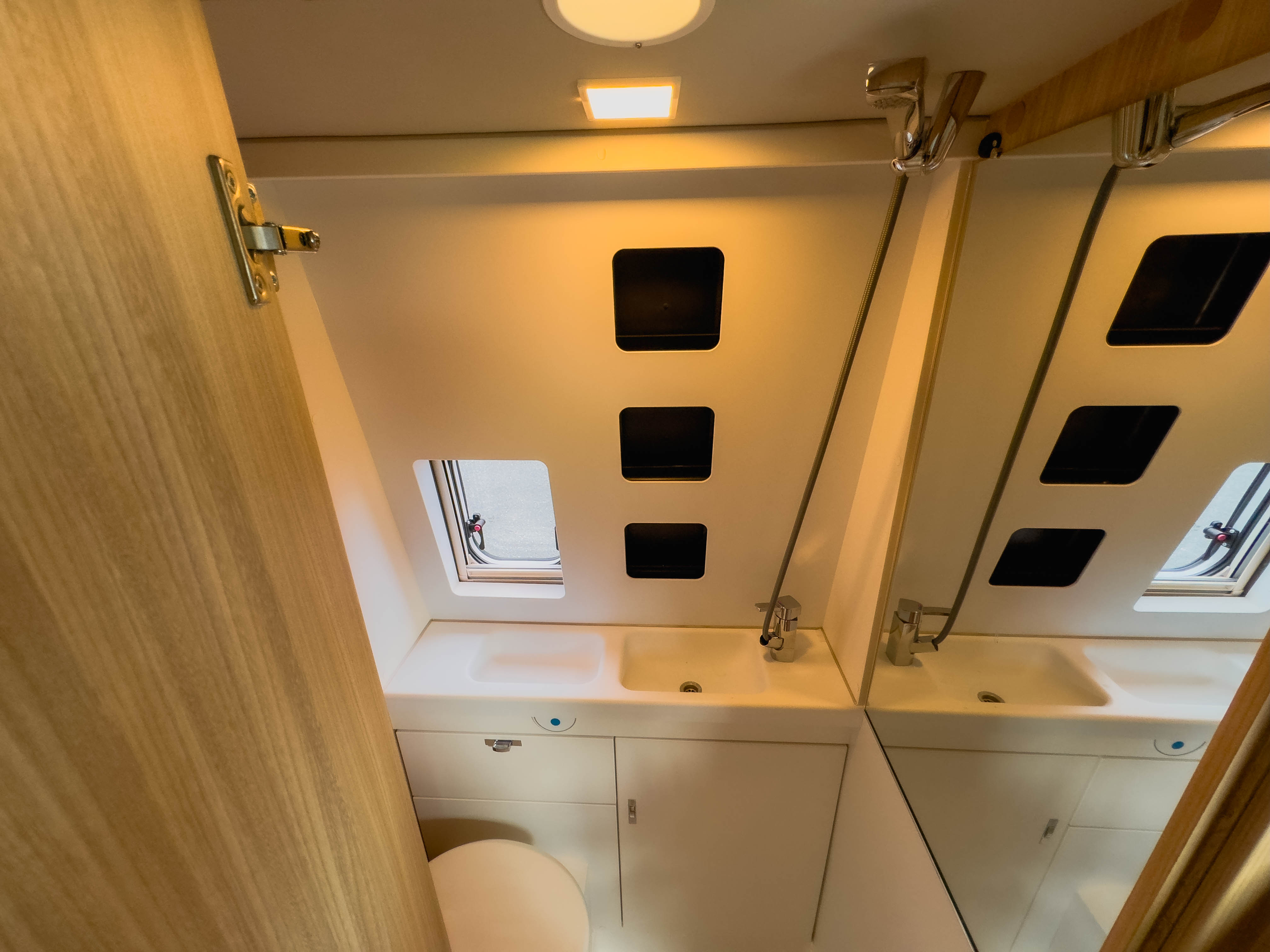 Campervan bathroom with sink, mirror, toilet, overhead light, and small window providing natural light and ventilation.