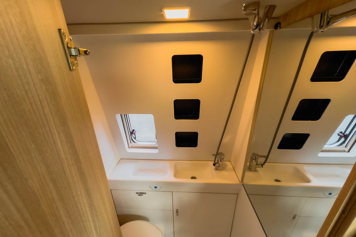 Campervan bathroom with sink, mirror, toilet, overhead light, and small window providing natural light and ventilation.