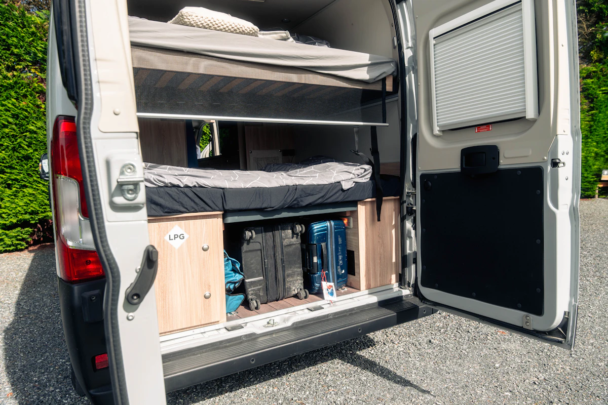 Rear view of campervan with open doors showing storage space, luggage, and a bed inside.