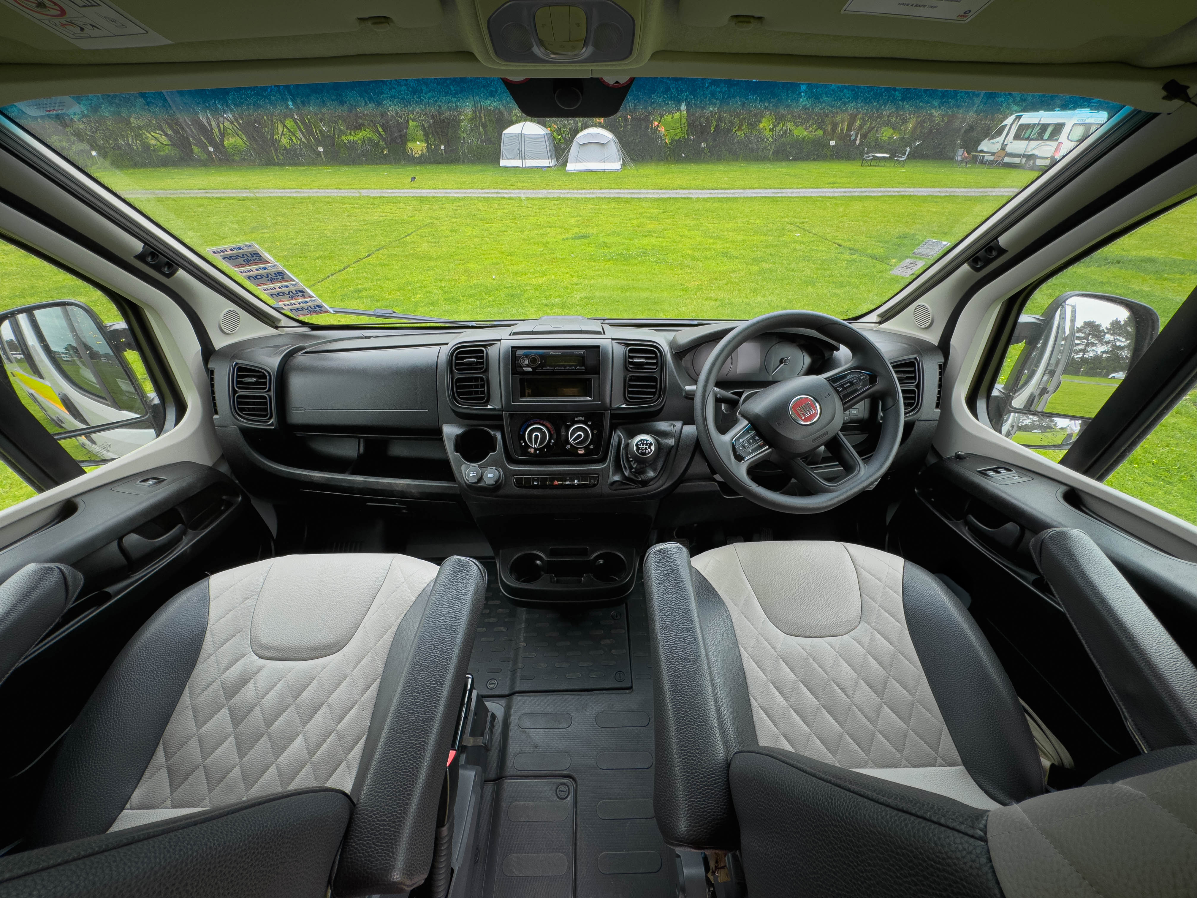 Campervan front interior with two seats, steering wheel, dashboard, and large windows overlooking a grassy campsite.