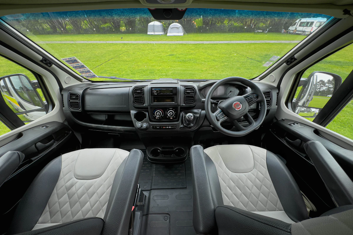 Campervan front interior with two seats, steering wheel, dashboard, and large windows overlooking a grassy campsite.