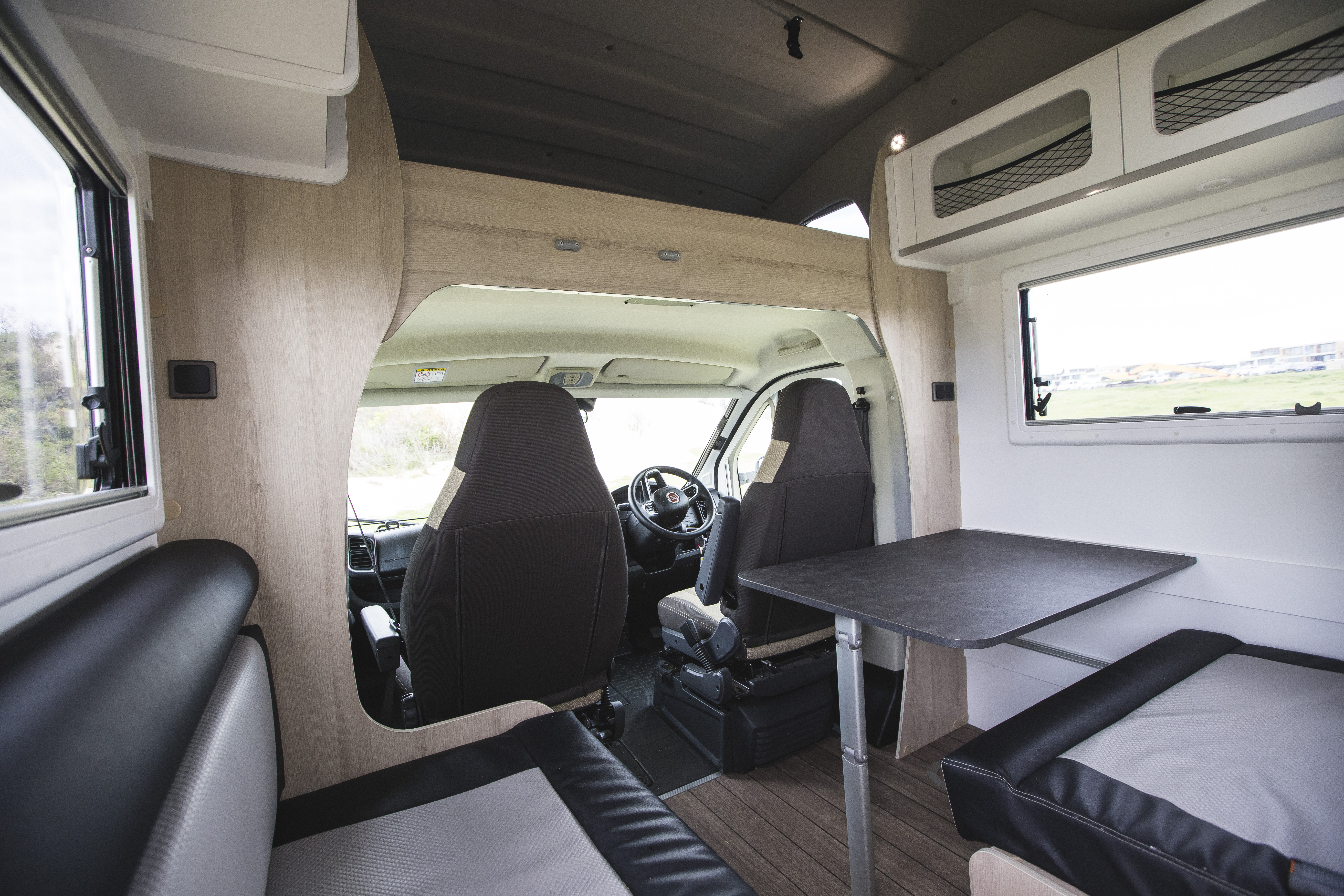 Modern campervan interior with two swivel front seats, dining table, bench seating, large windows, and overhead storage.