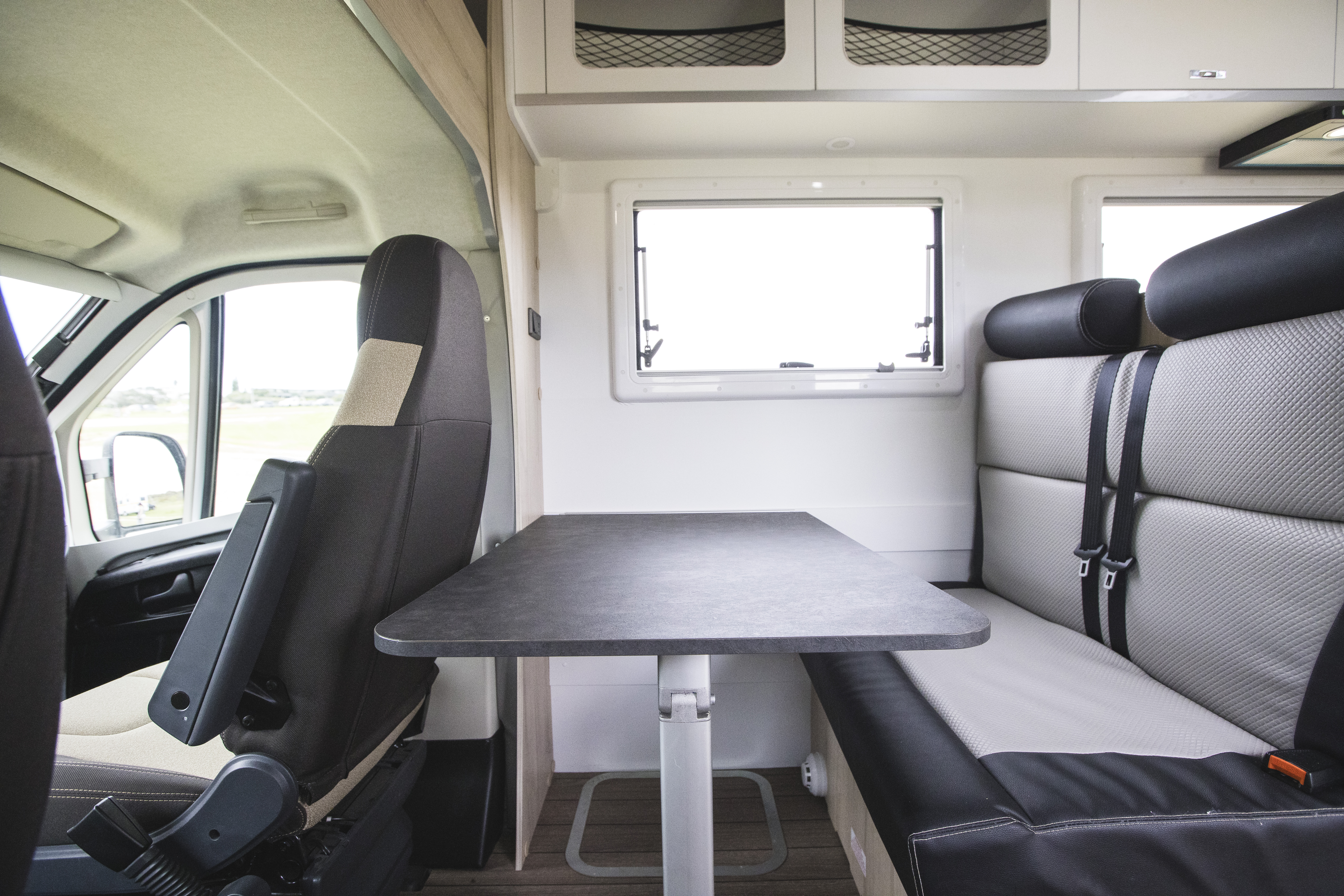 Modern campervan interior with a dining table, front driver seat, cushioned bench seats, and large windows.
