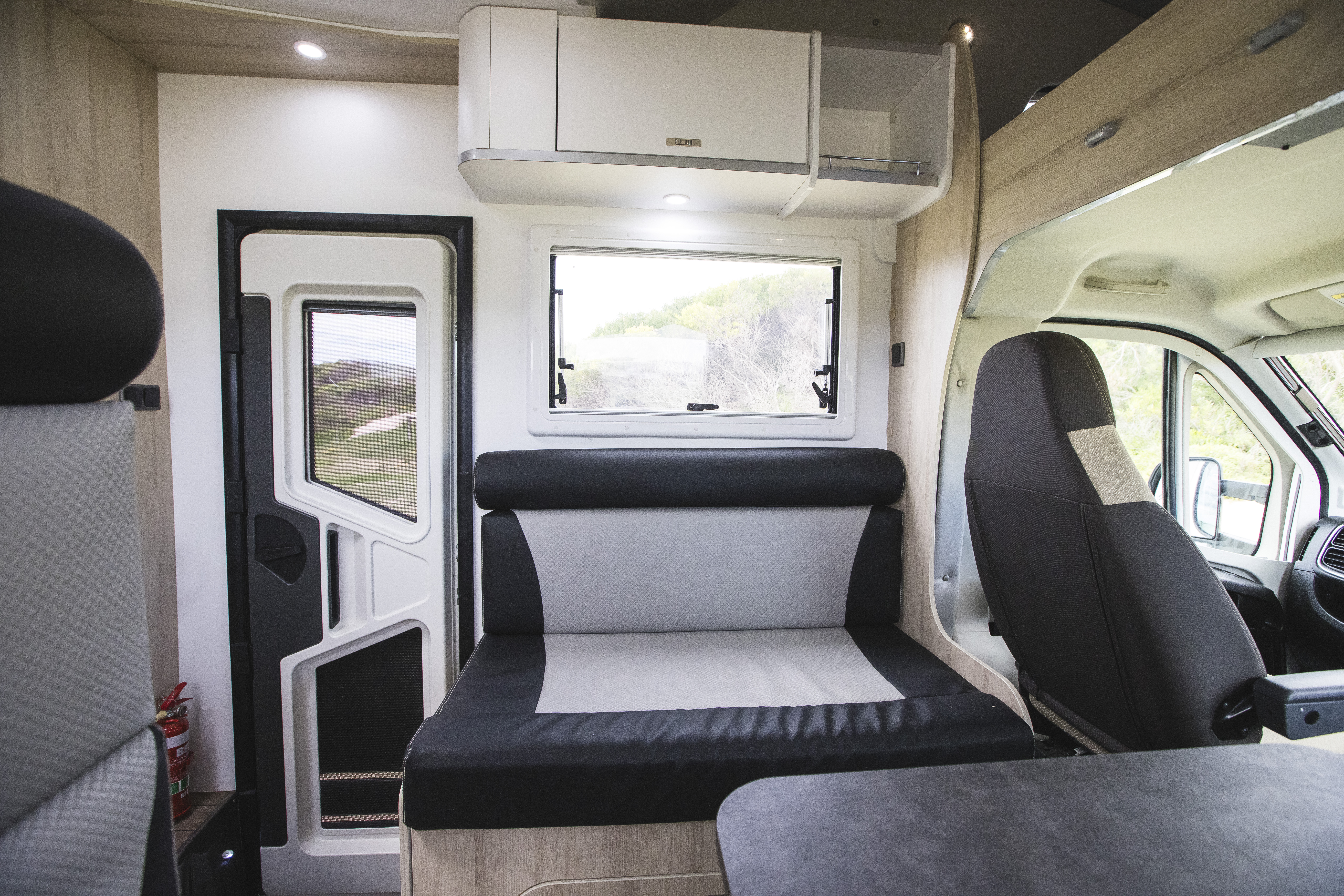 Modern campervan interior with seating area, driver’s seat, window, overhead storage, and door, in natural daylight.