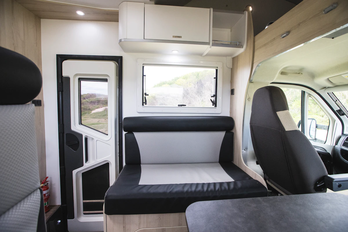 Modern campervan interior with seating area, driver’s seat, window, overhead storage, and door, in natural daylight.
