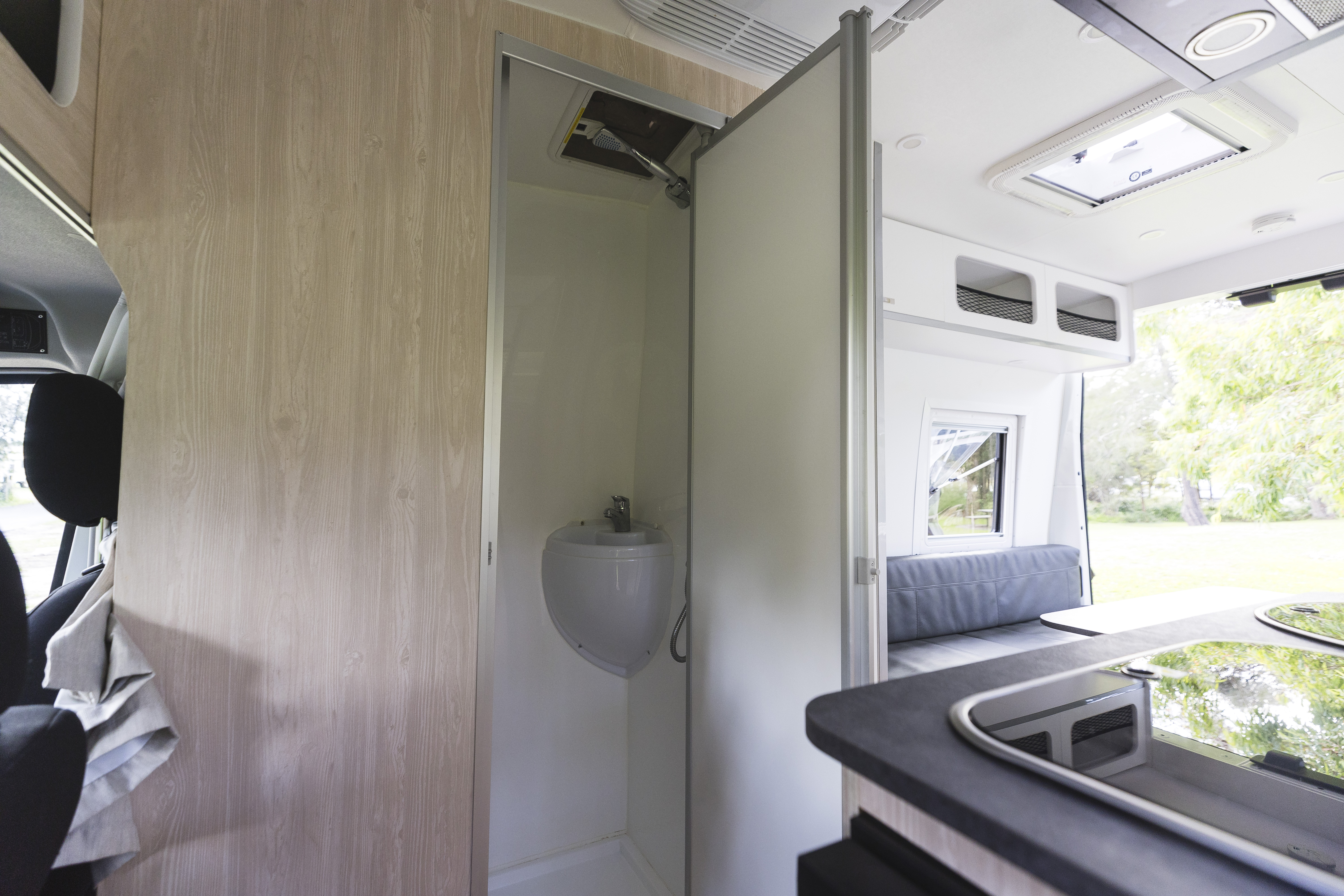 Interior of a campervan showing a compact bathroom with toilet, kitchen counter, and seating area by a window.