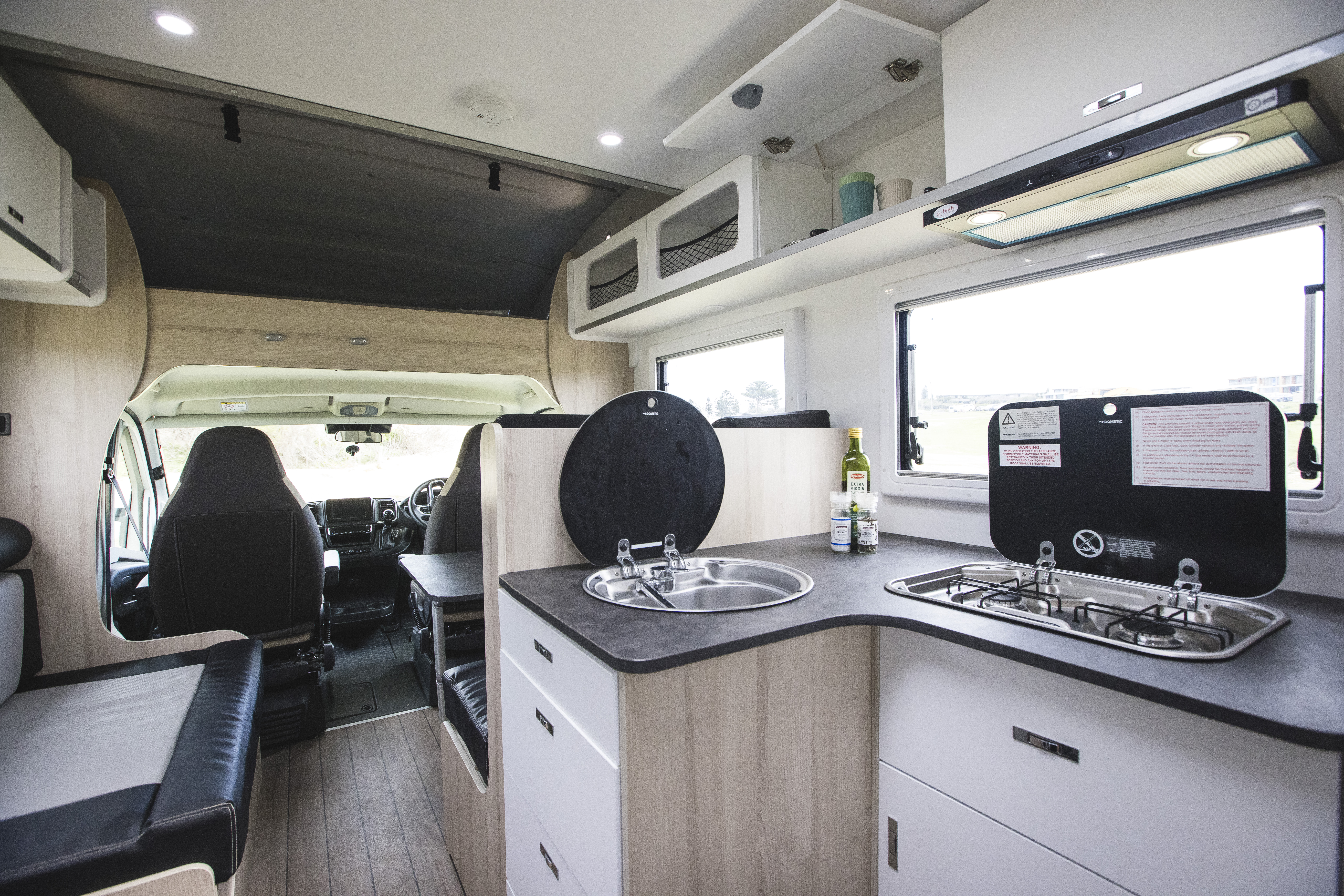 Modern campervan interior with kitchen, sink, stovetop, seating area, storage cabinets, and front driver and passenger seats.