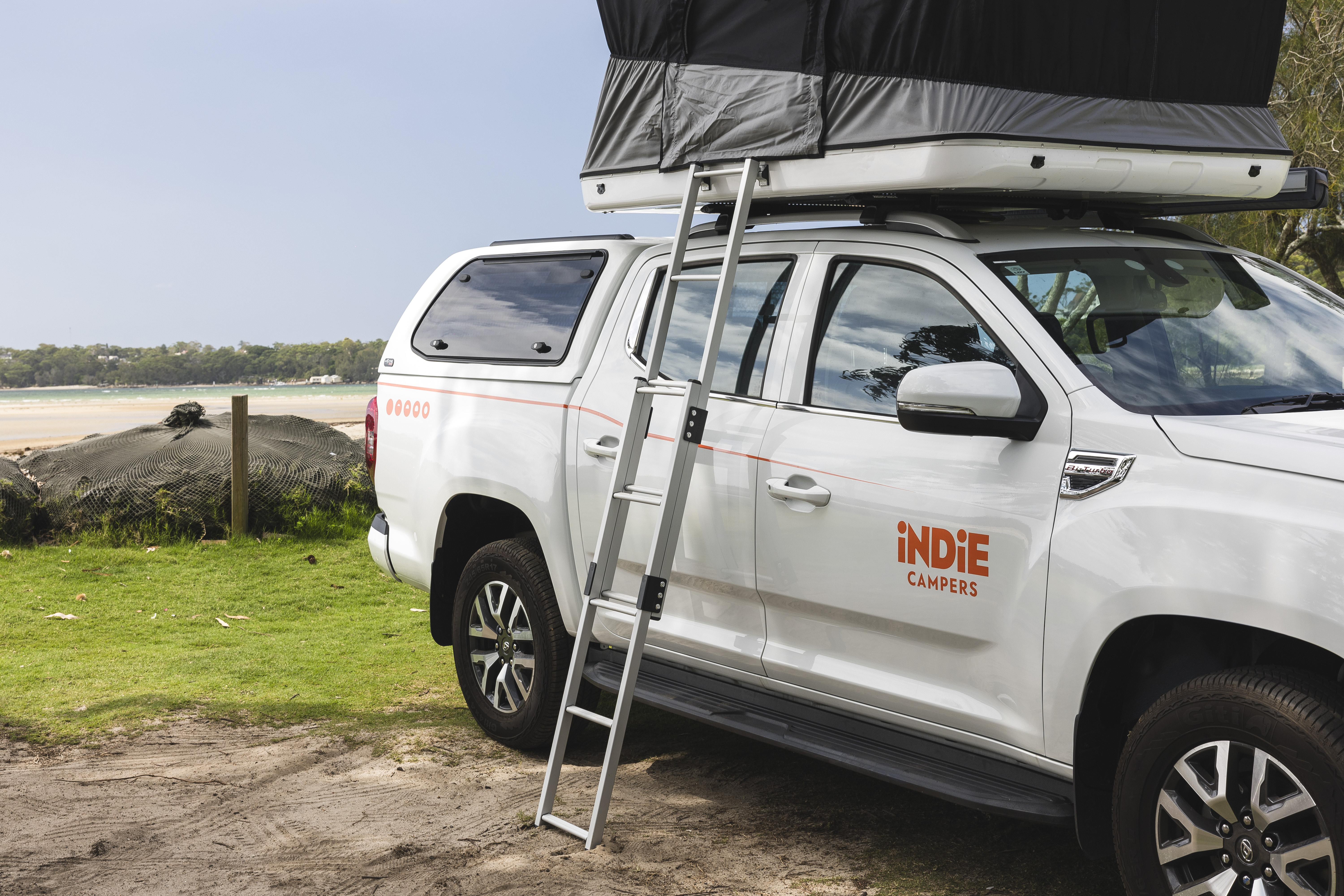 White campervan with rooftop tent and ladder, parked on grass near the beach, Indie Campers logo visible.