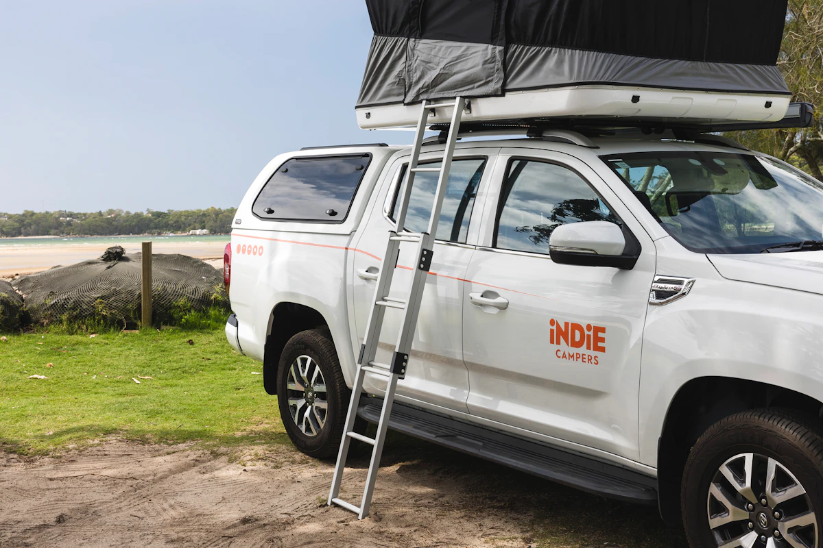 White camper van with rooftop tent and ladder, parked on grass near the beach, Indie Campers logo visible.