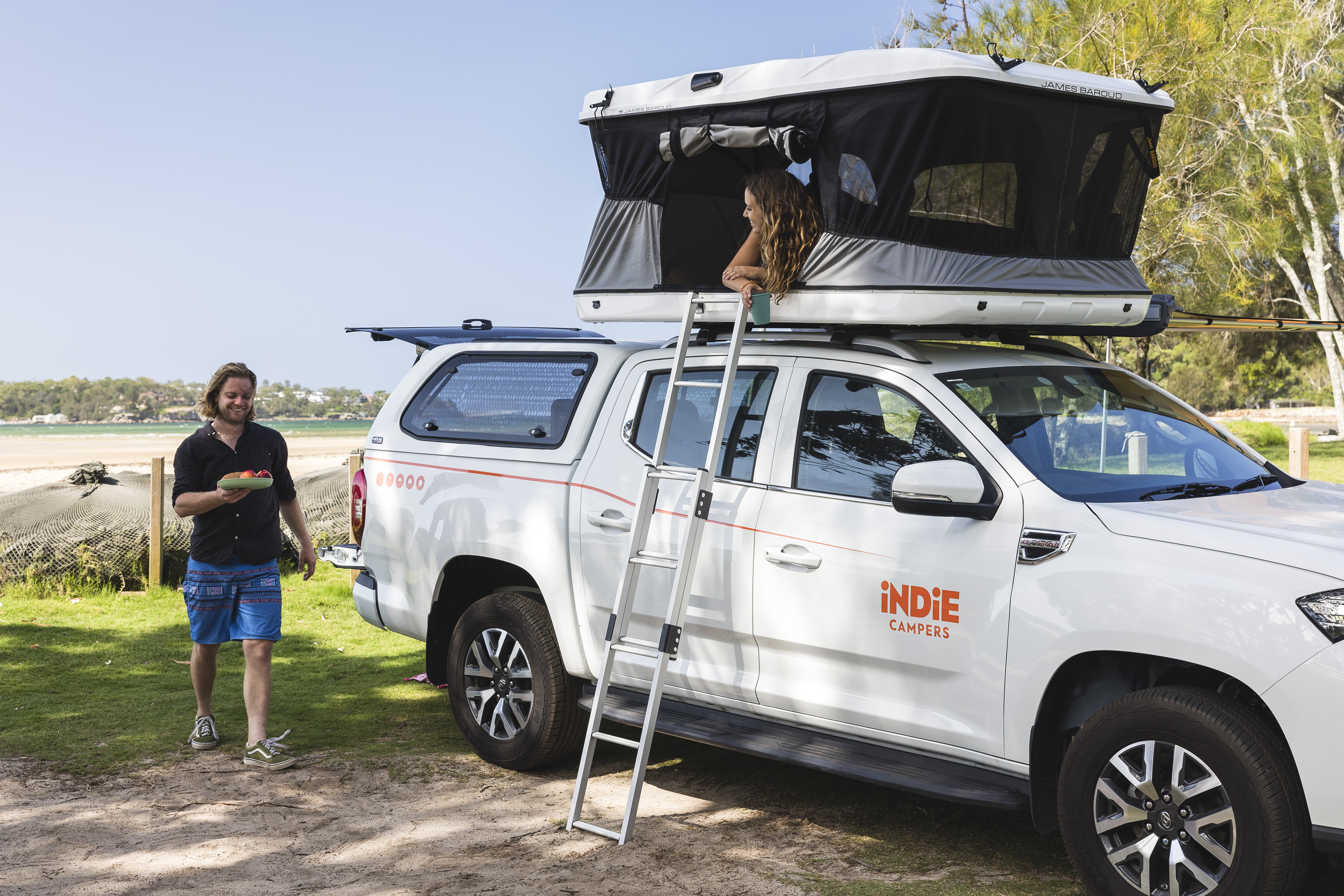 Man holding food near Indie Campers van with rooftop tent, woman sitting inside tent, outdoor nature setting.