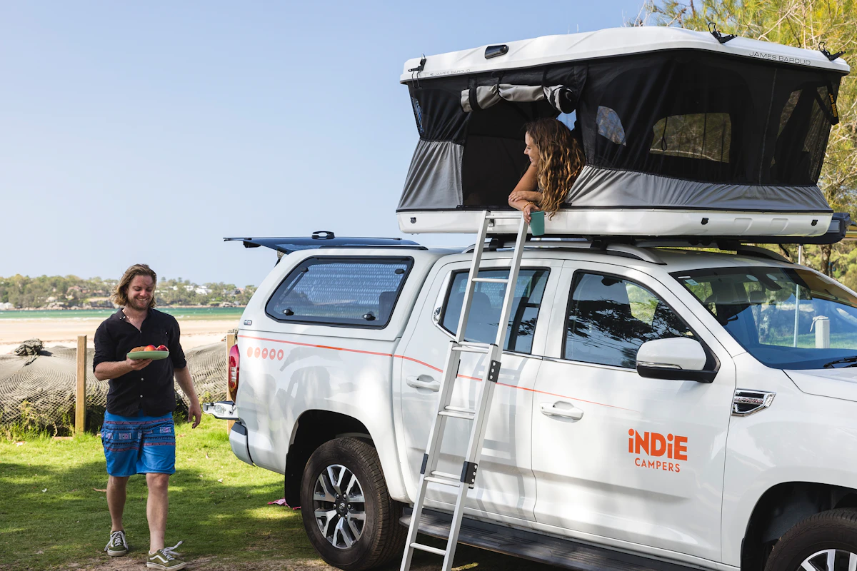 Man holding food near Indie Campers van with rooftop tent, woman sitting inside tent, outdoor nature setting.
