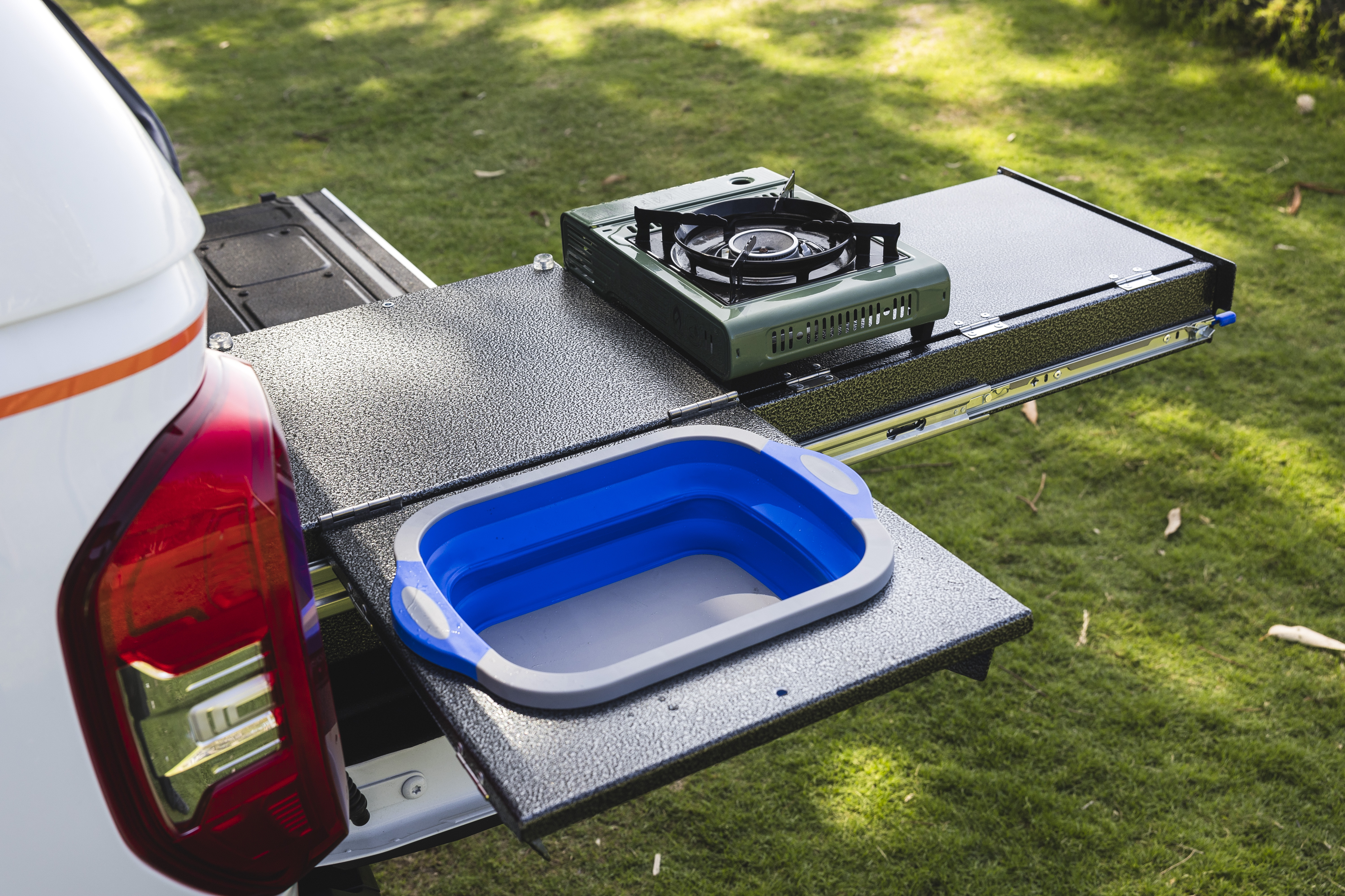 Campervan outdoor kitchen setup with portable gas stove and collapsible sink on pull-out trays, parked on grass.