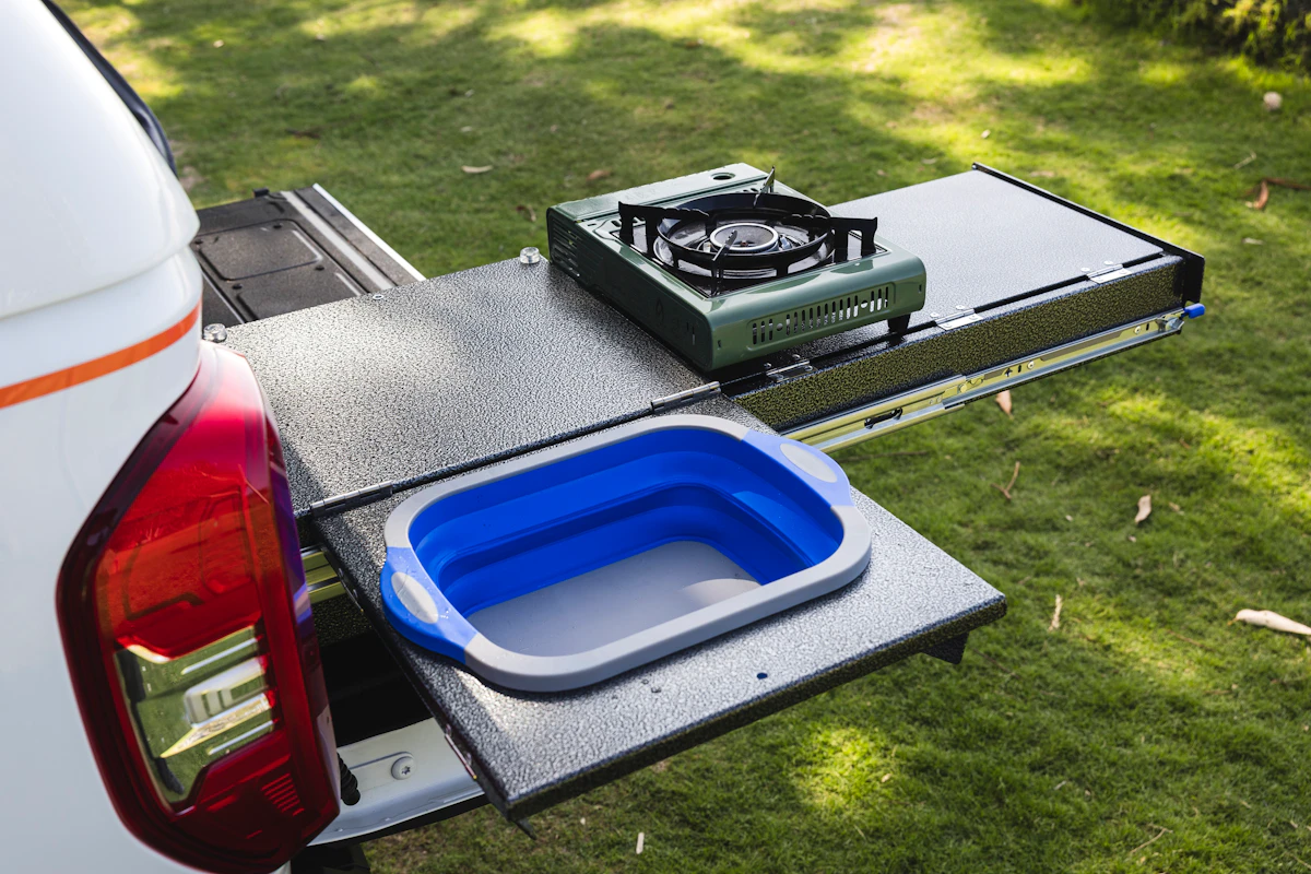 Campervan outdoor kitchen setup with portable gas stove and collapsible sink on pull-out trays, parked on grass.