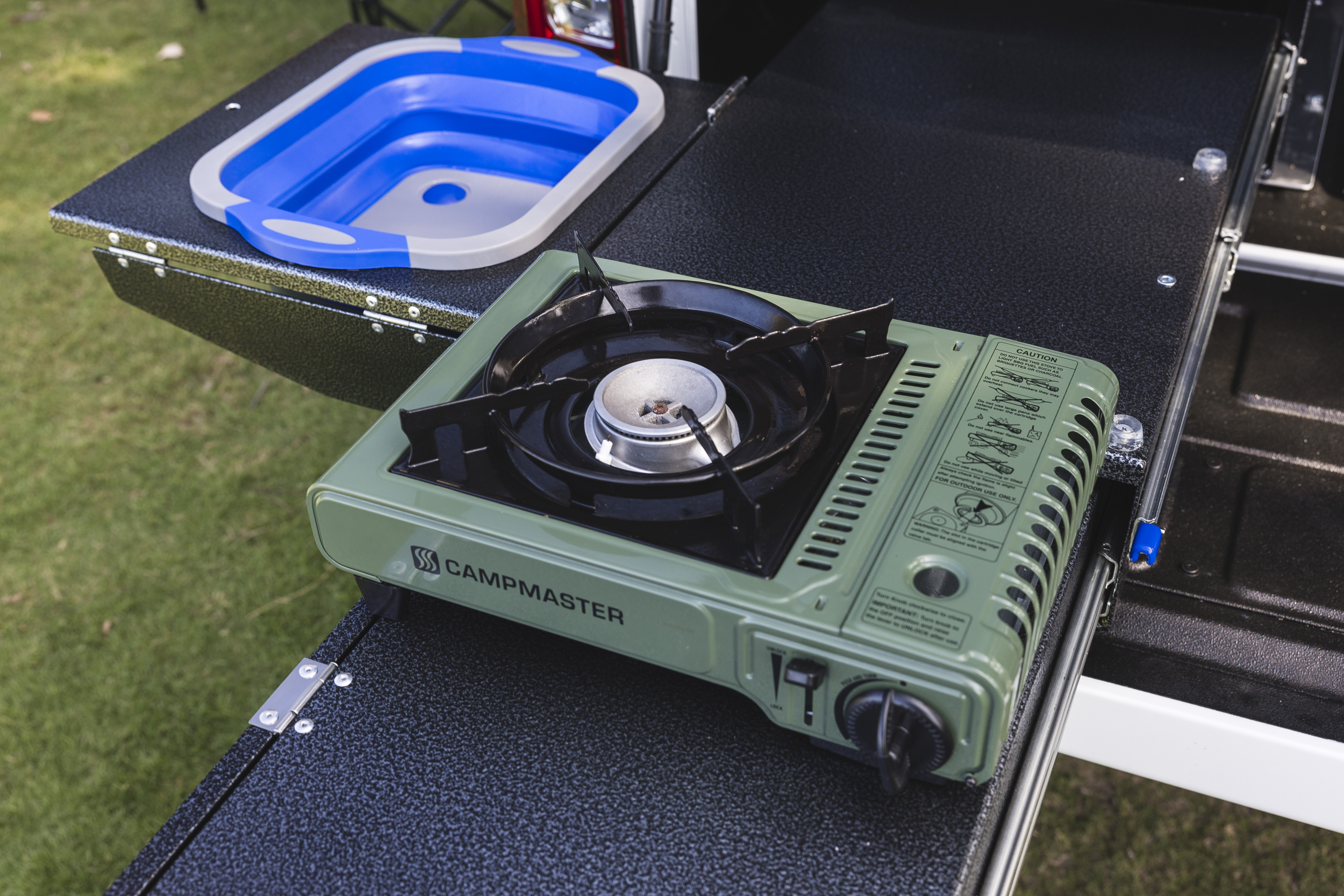 Portable green gas stove and collapsible blue sink on a campervan outdoor kitchen setup, placed on a grassy area.