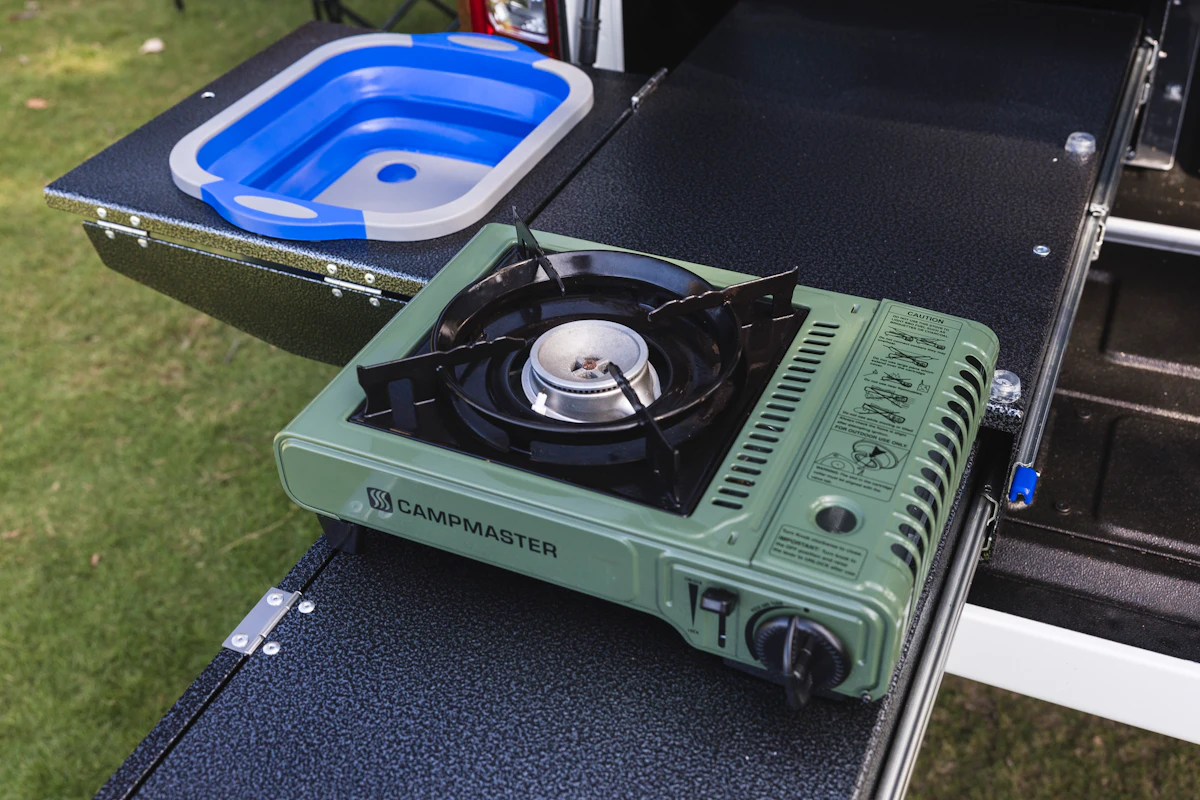Portable green gas stove and collapsible blue sink on a campervan outdoor kitchen setup, placed on a grassy area.