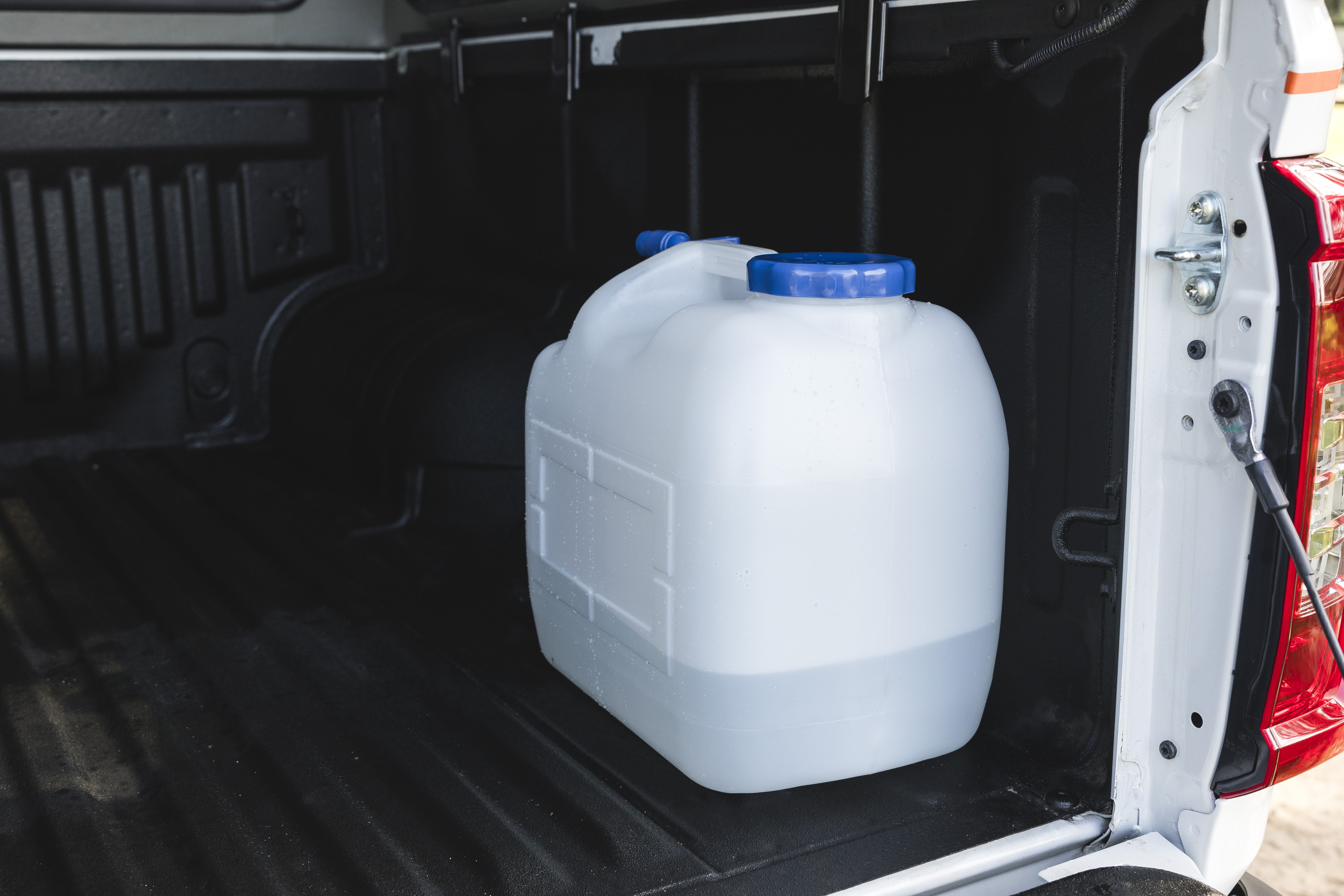 Large plastic water container with blue cap placed in the back of an open campervan or vehicle.
