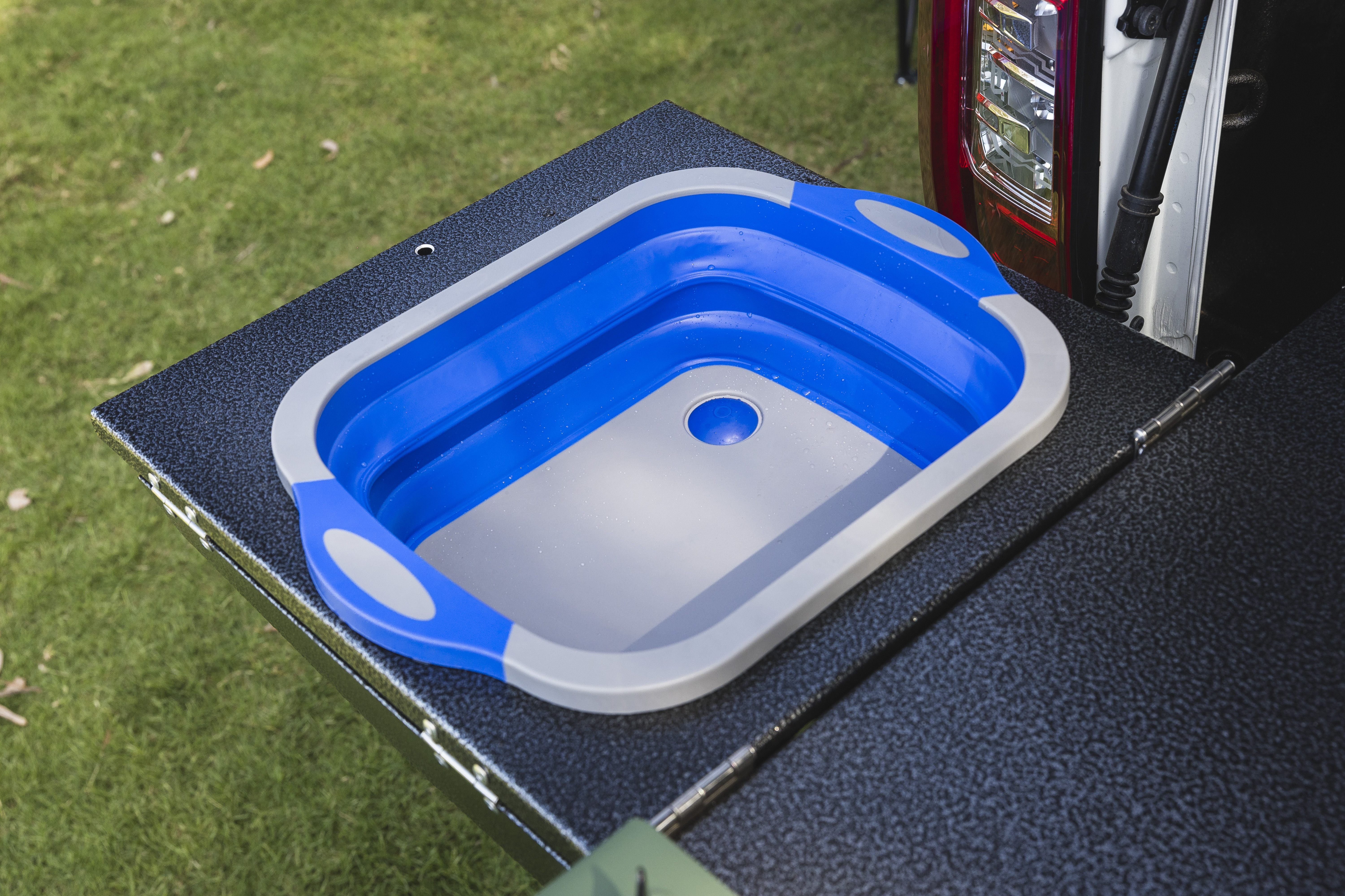 Collapsible blue and grey sink set up on a campervan’s outdoor kitchen countertop with grass in background.