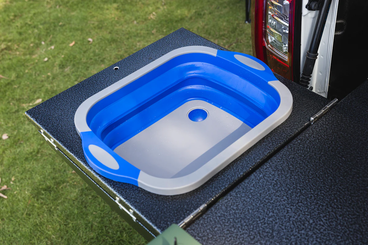 Collapsible blue and gray sink set up on a campervan’s outdoor kitchen countertop with grass in background.