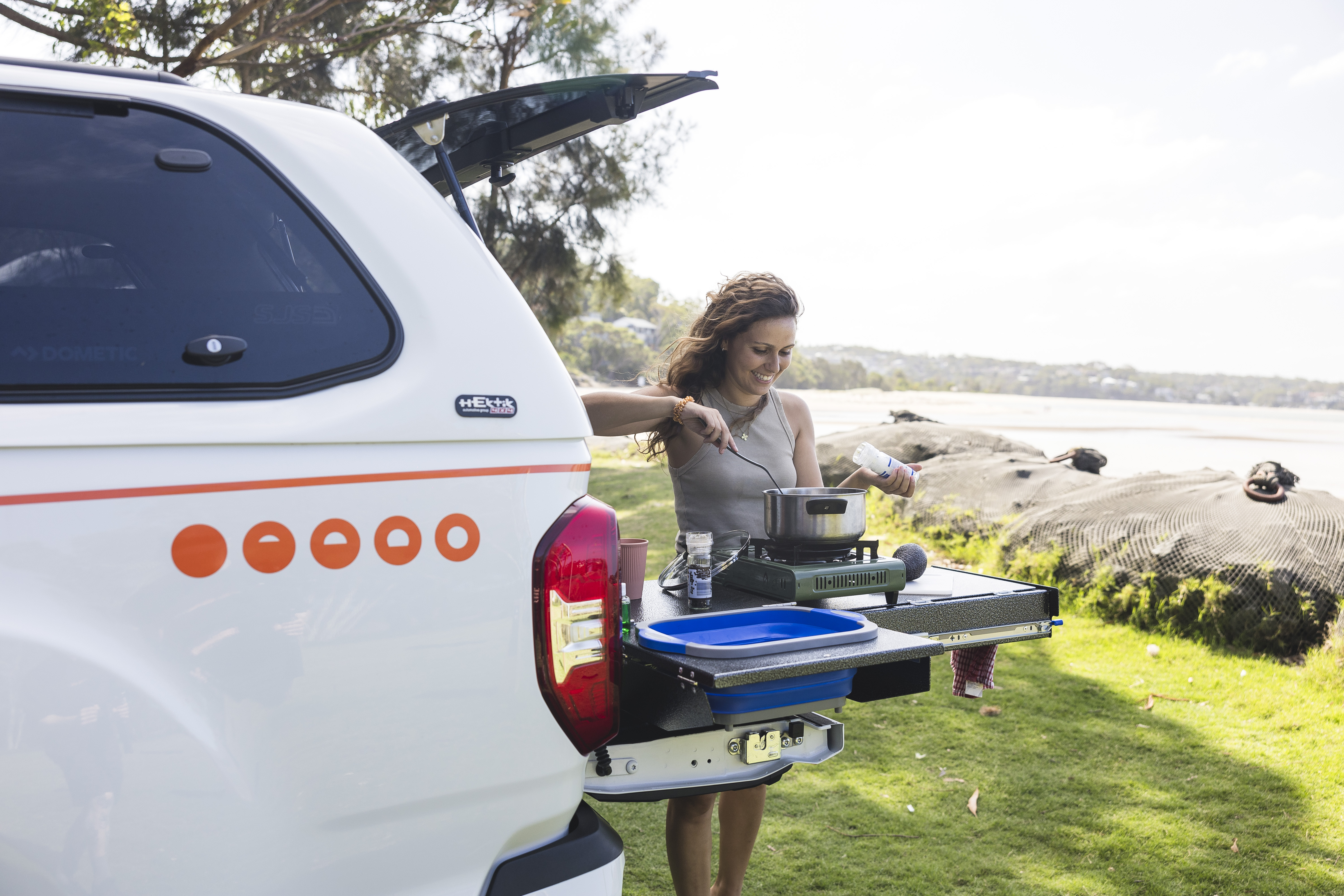 Woman cooking on outdoor kitchen setup attached to campervan by a scenic lakeside location under clear sky.