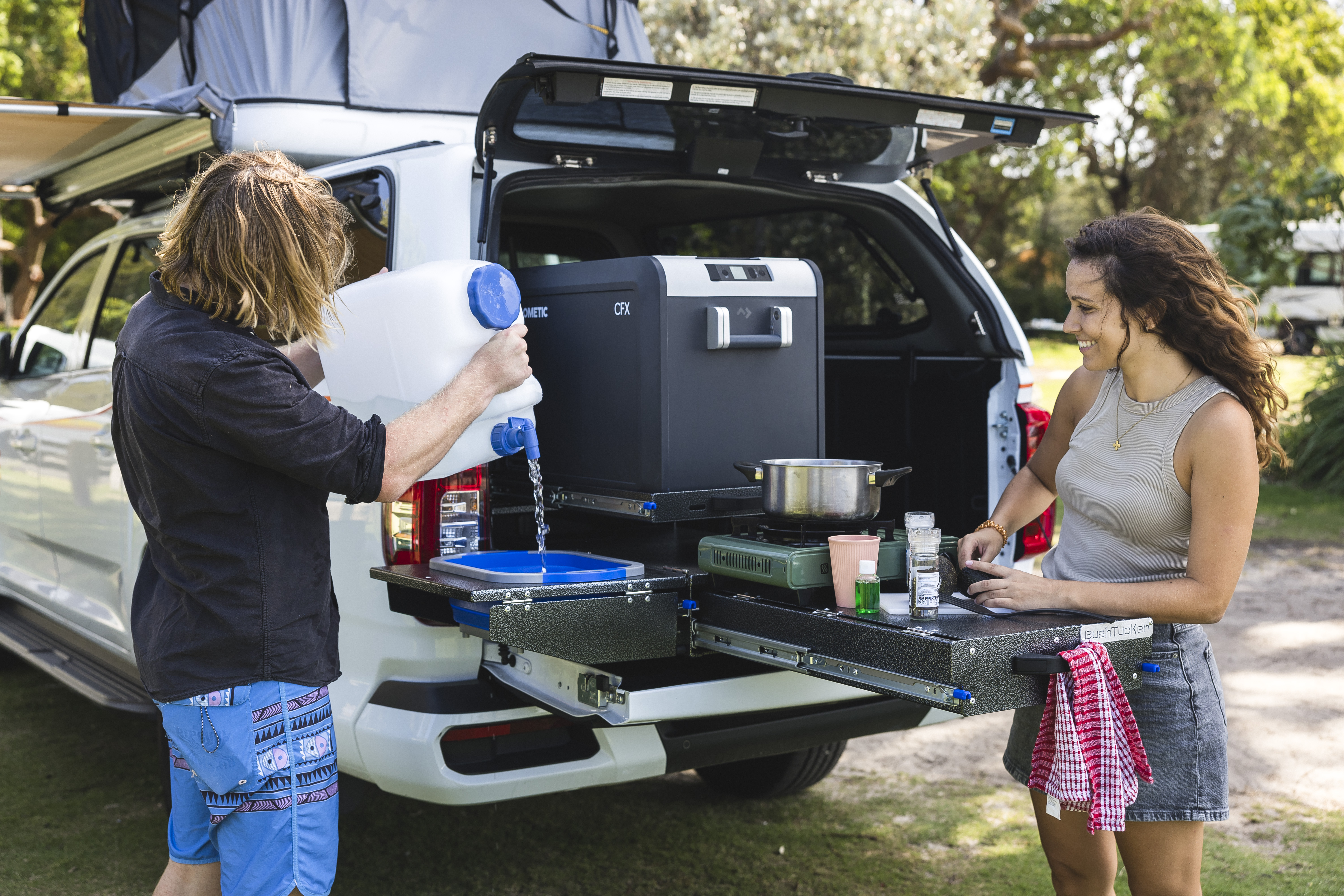 Two people cooking at a pull-out kitchen in the back of a campervan parked outdoors.