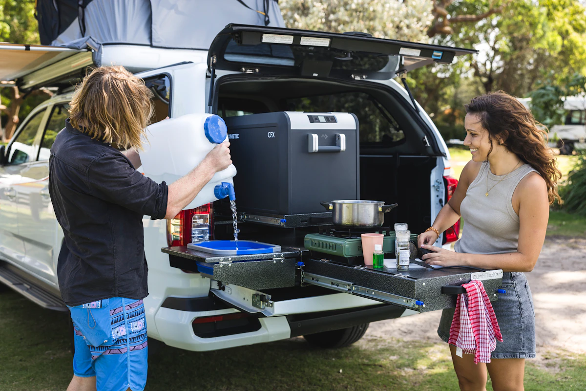 Two people cooking at a pull-out kitchen in the back of a camper van parked outdoors.