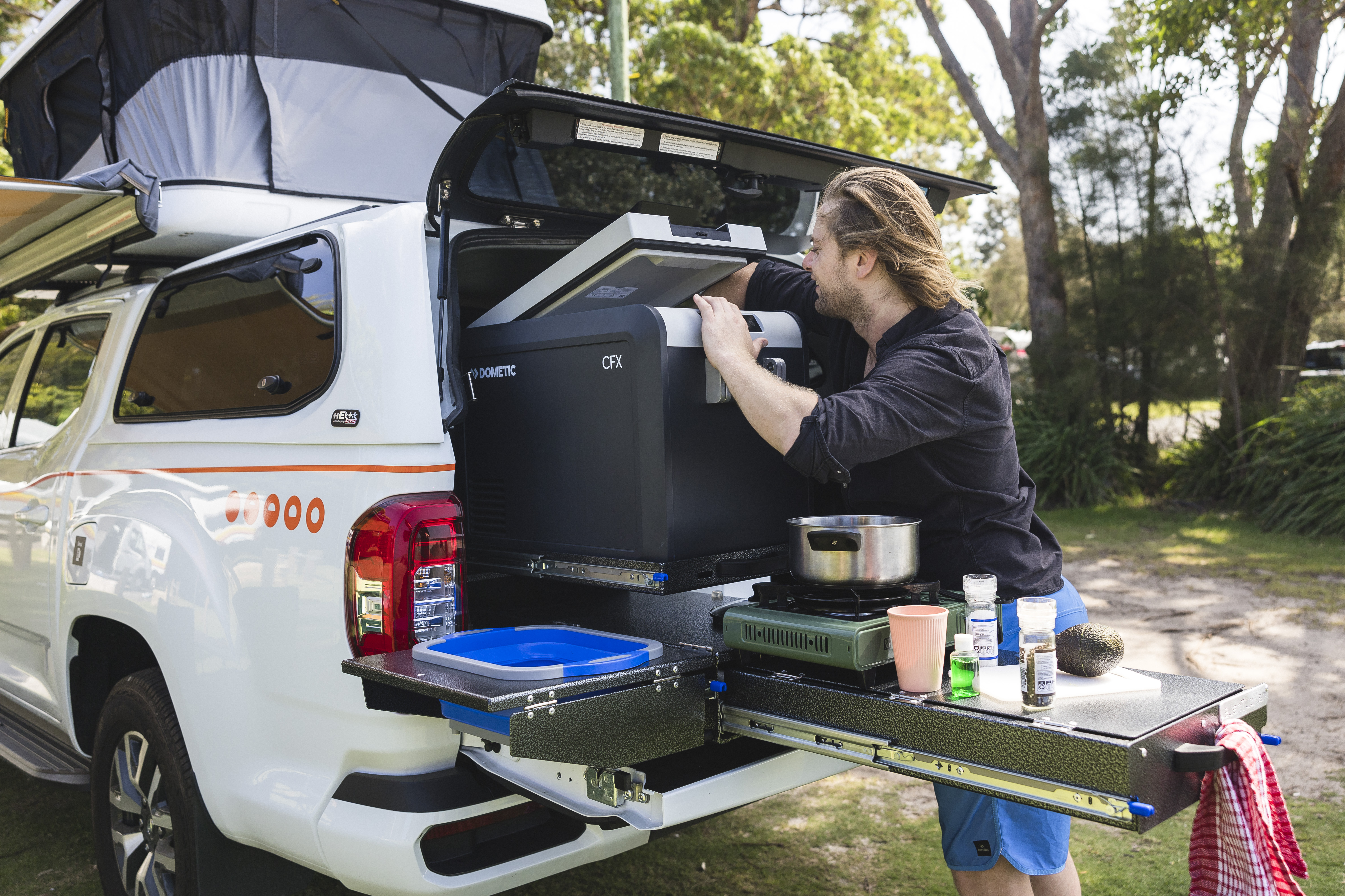 Person preparing food at a pull-out kitchen in the back of a white campervan at a campsite.
