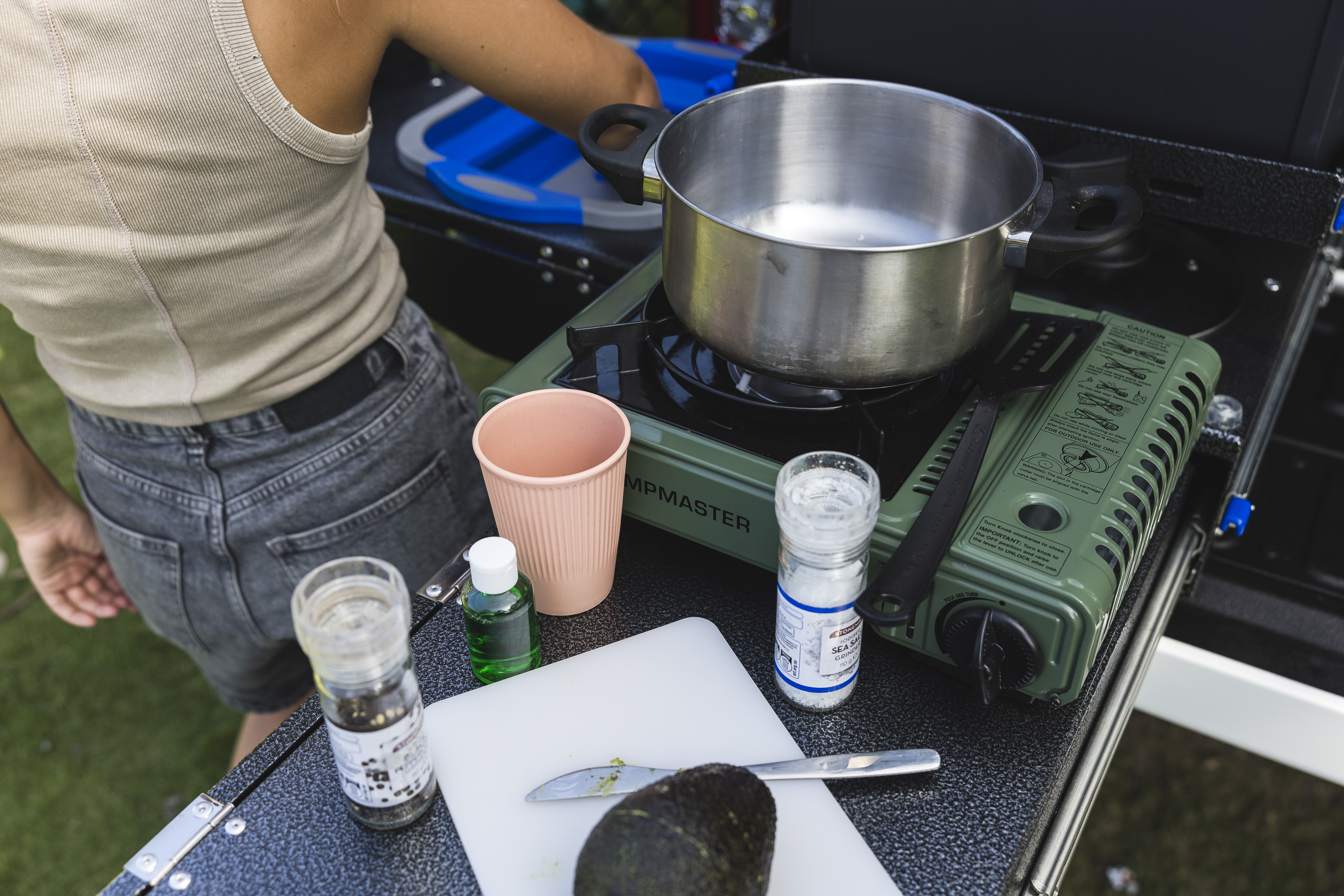 Outdoor camper kitchen setup with portable stove, cookware, spices, a cutting board, and a person preparing food.