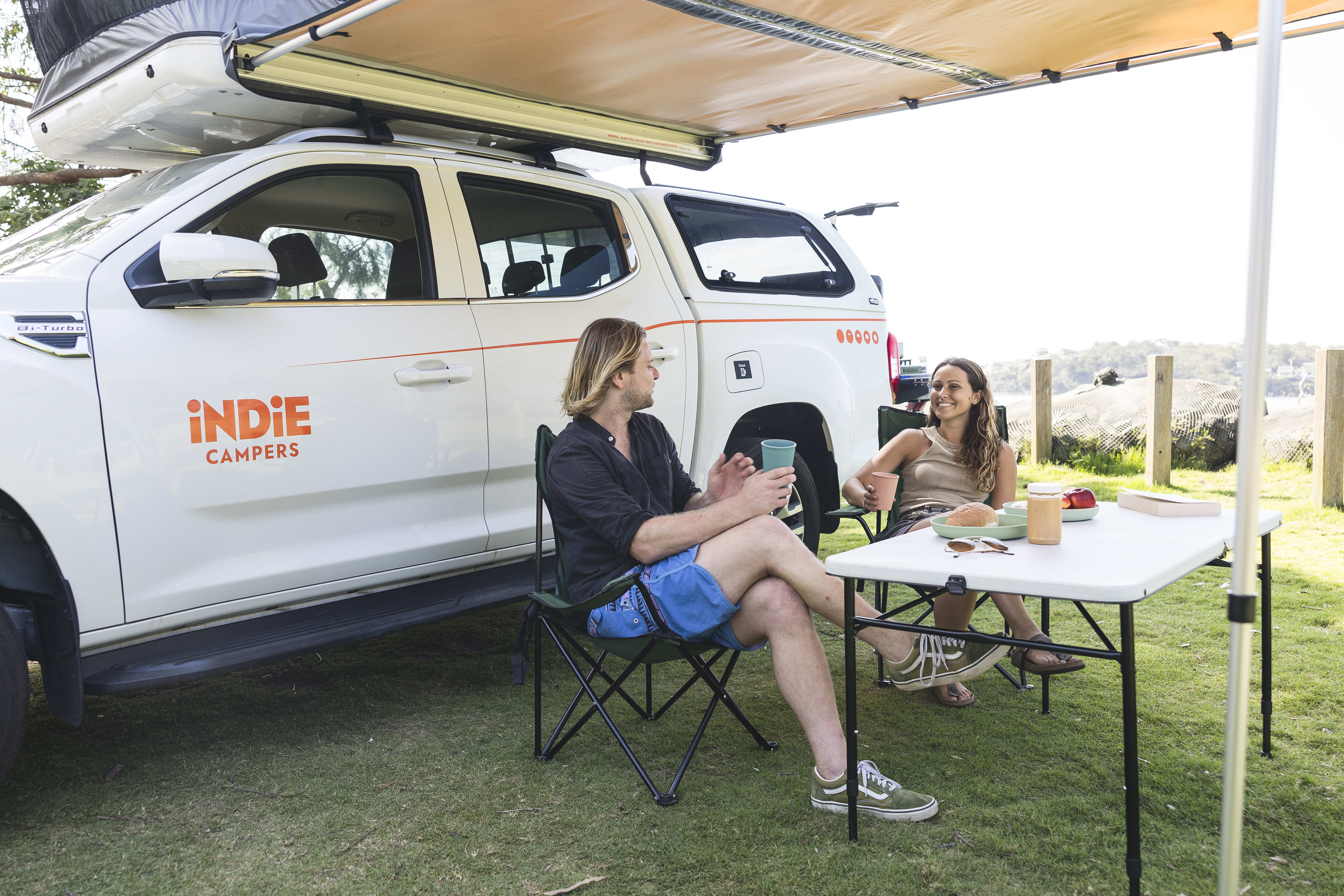 Two people relaxing and enjoying drinks at a campsite beside an Indie Campers vehicle on a sunny day.
