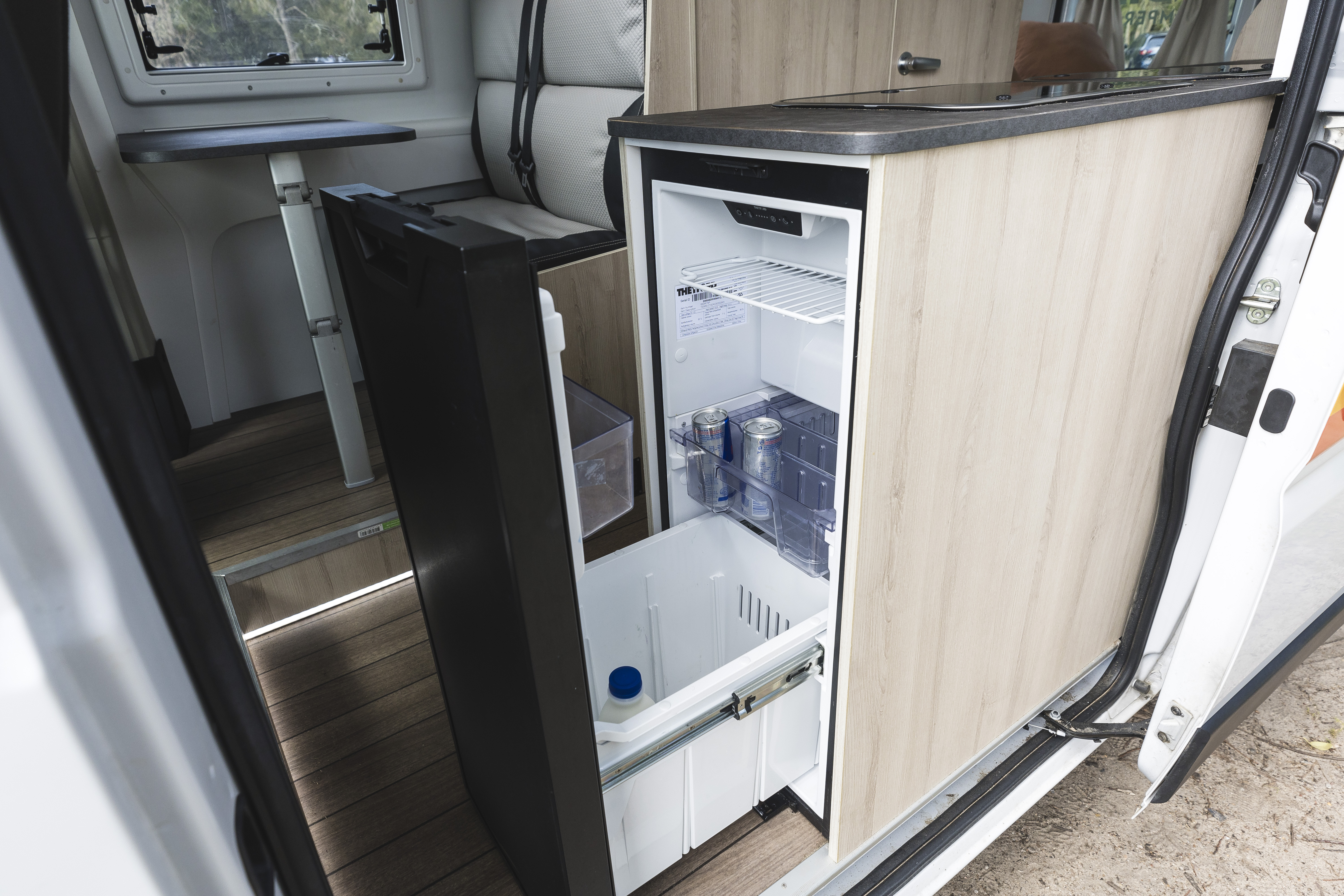 Compact camper van refrigerator and freezer open, showing drinks and storage inside, with modern wood-finish kitchenette.