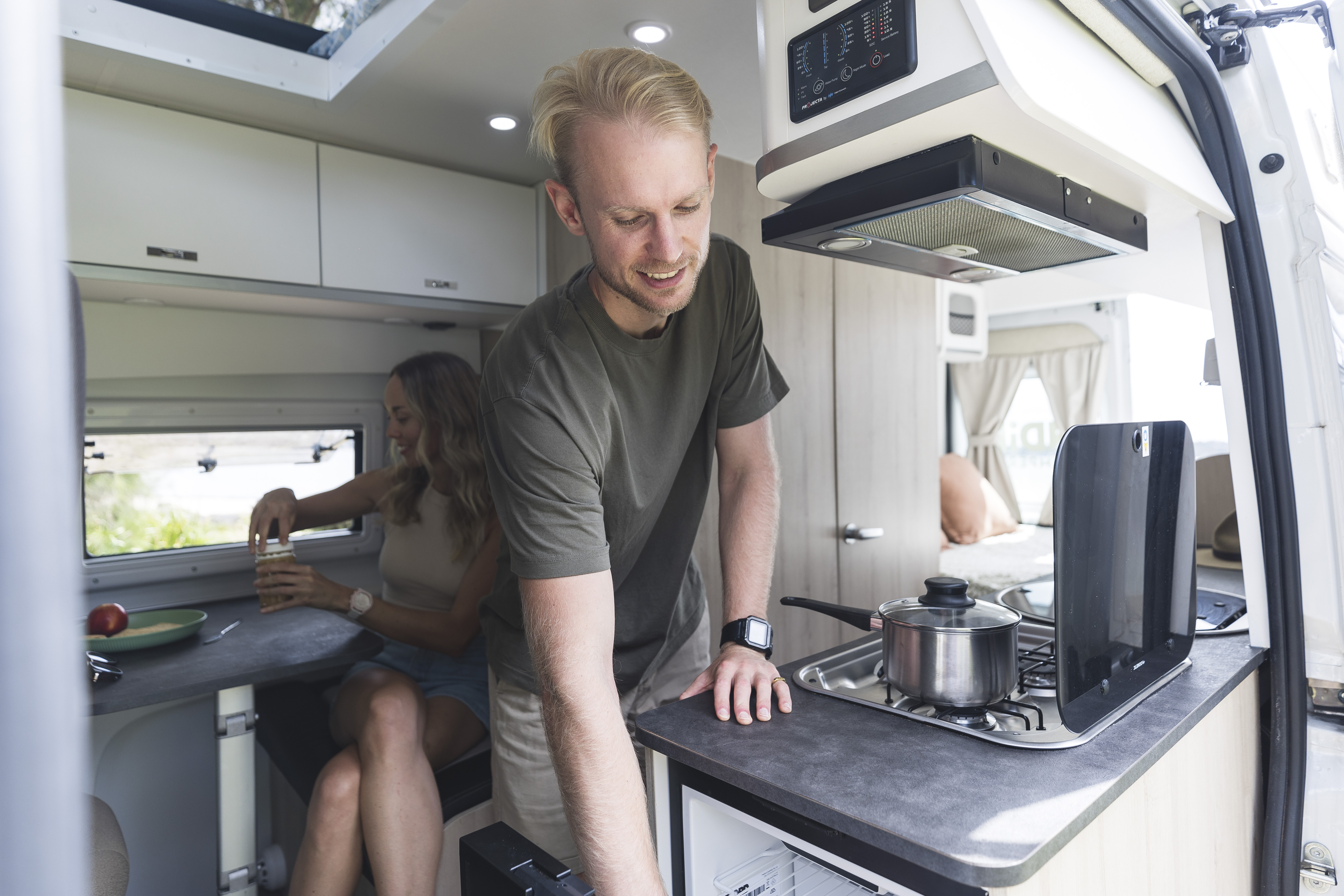 Man cooking in a modern camper van kitchen while a woman sits at the dining area inside the vehicle.
