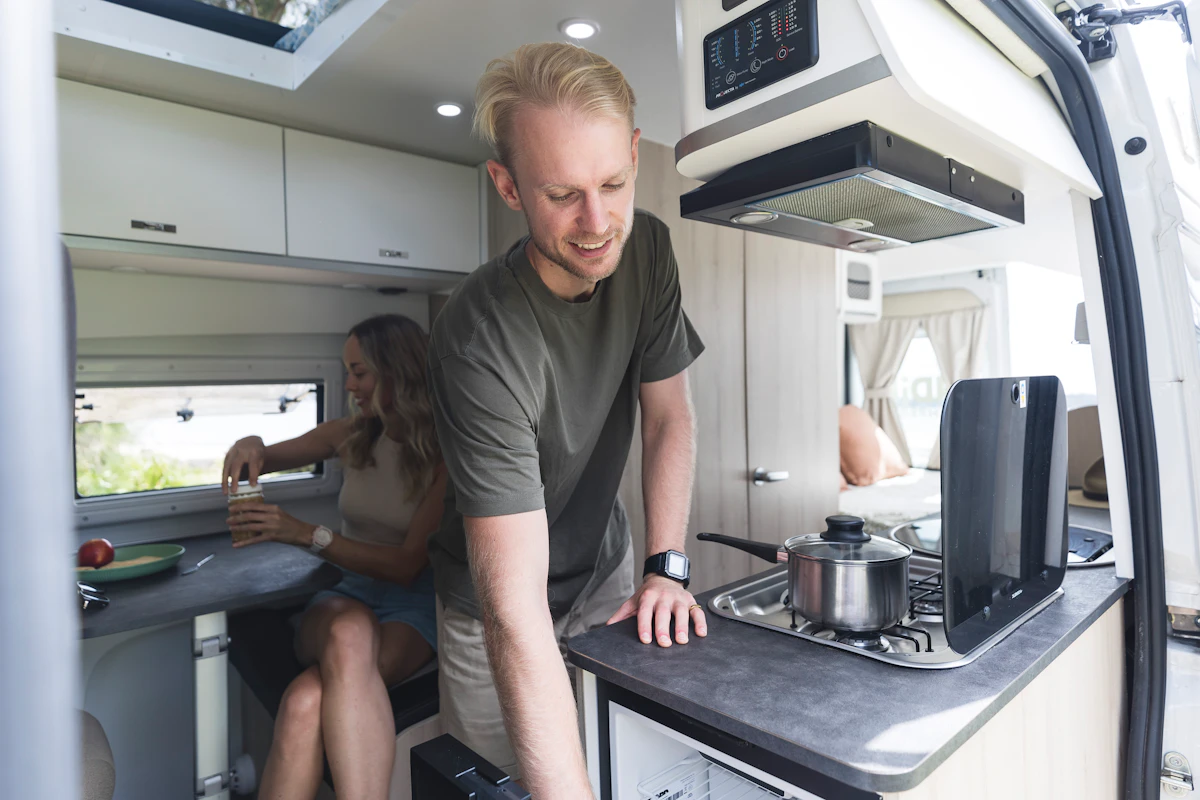Man cooking in a modern camper van kitchen while a woman sits at the dining area inside the vehicle.