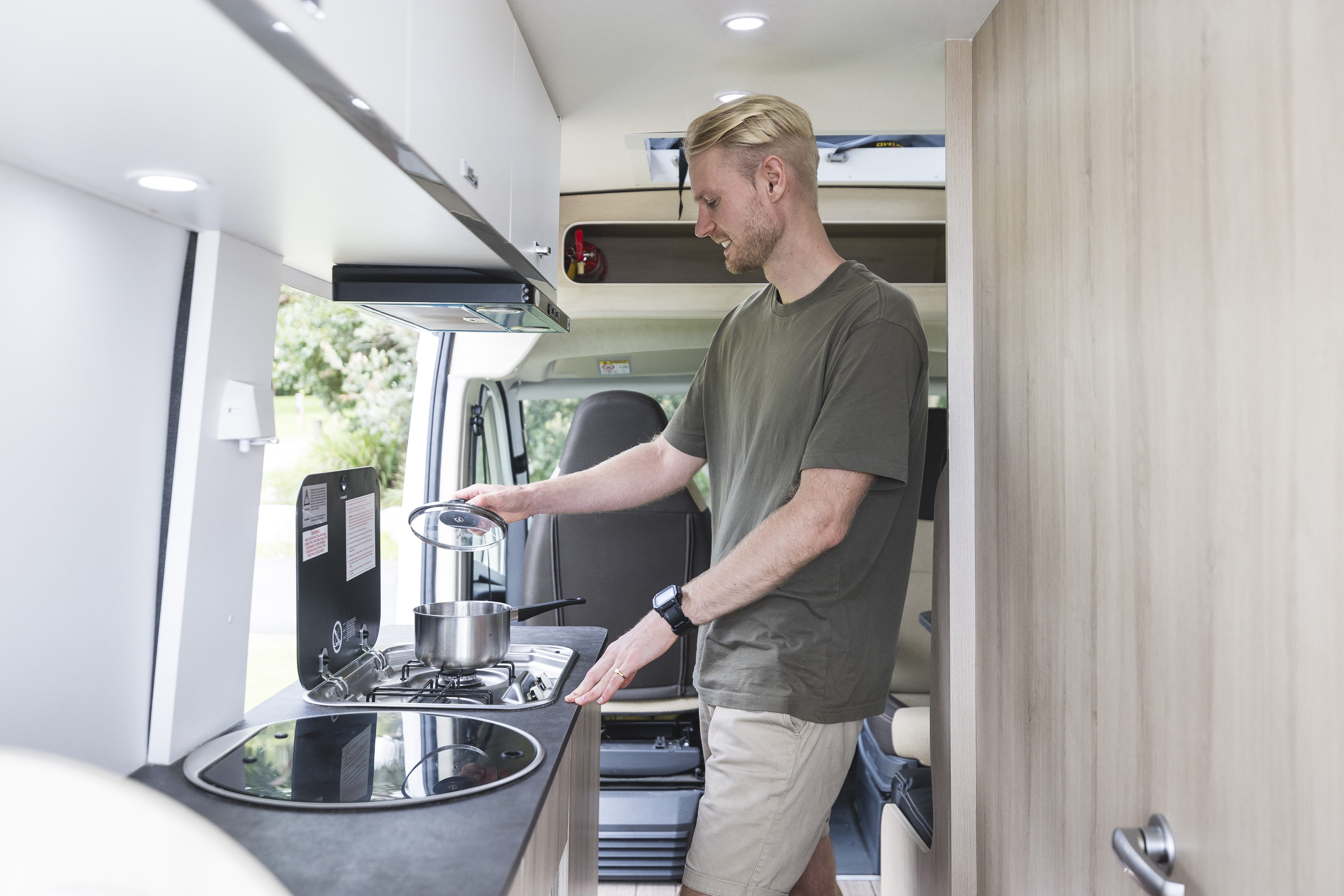 Person cooking with a pot on a stove inside a modern camper van kitchen during the day.