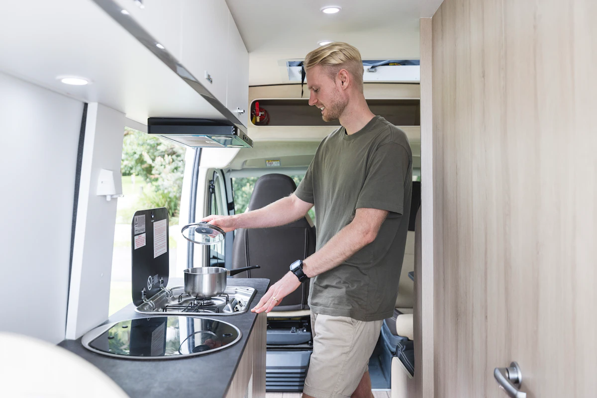 Person cooking with a pot on a stove inside a modern campervan kitchen during the day.