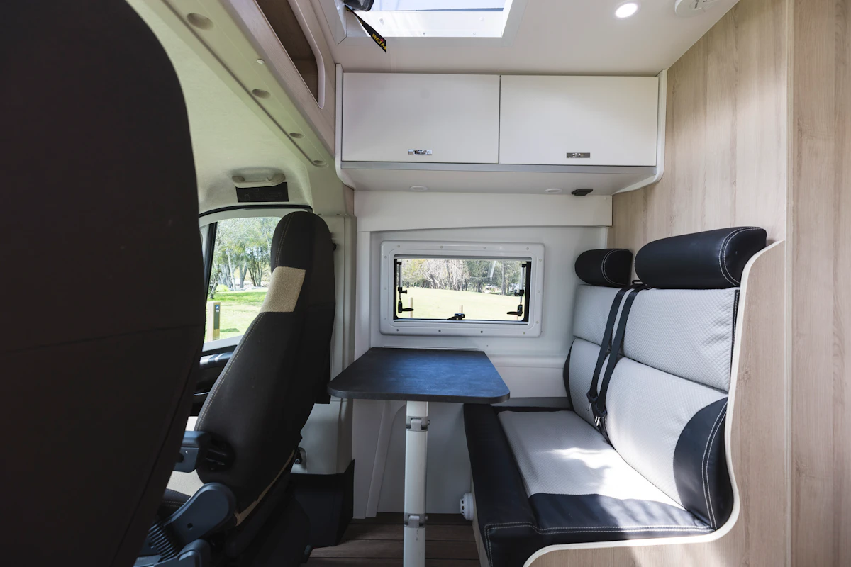 Modern campervan interior with swivel driver seat, dining table, bench seating, window, and overhead storage cabinets.