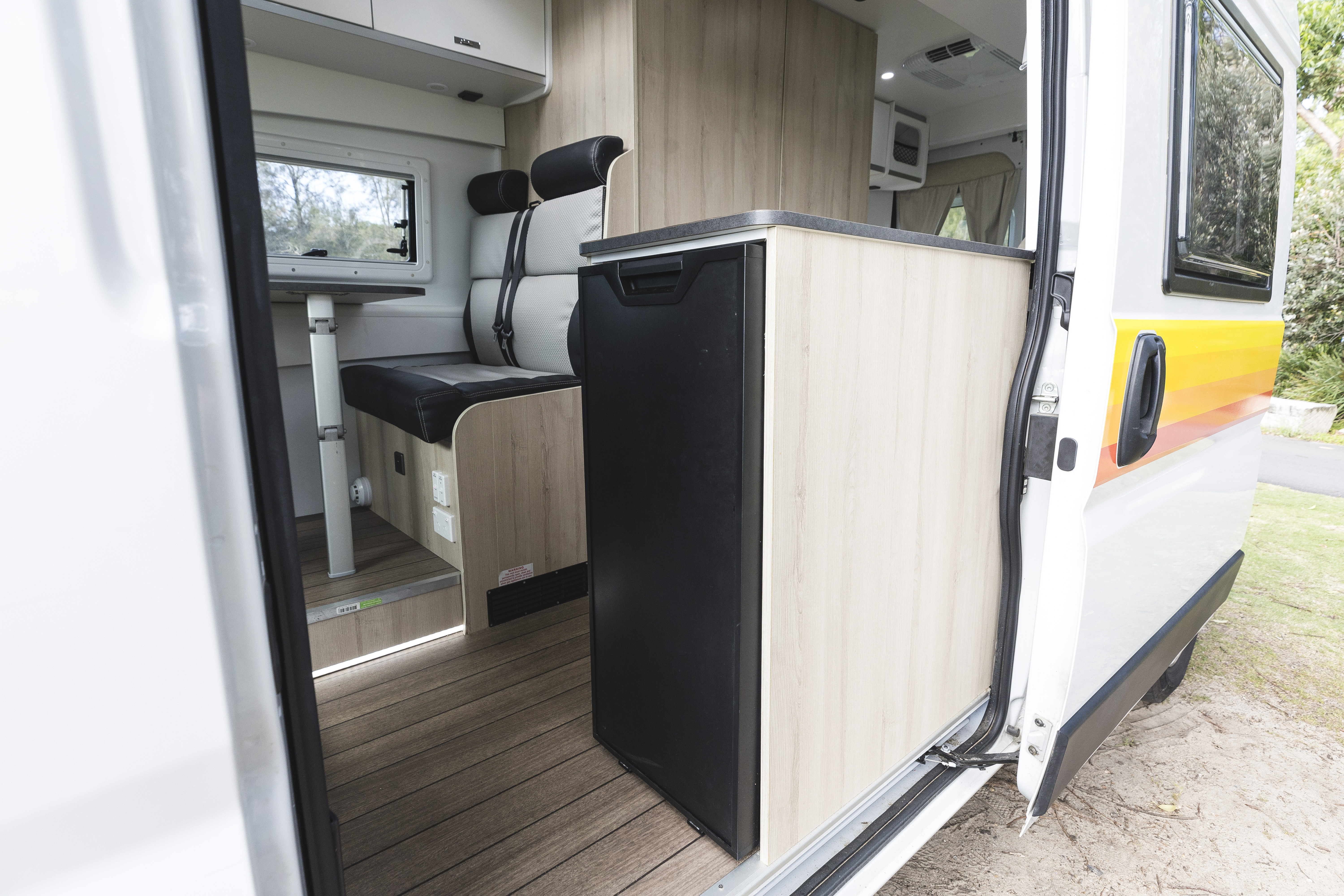 Interior view of campervan showing entrance, kitchenette with fridge, and seating area with table beside a window.