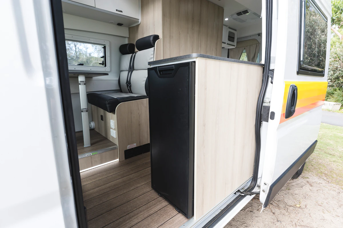 Interior view of campervan showing entrance, kitchenette with fridge, and seating area with table beside a window.