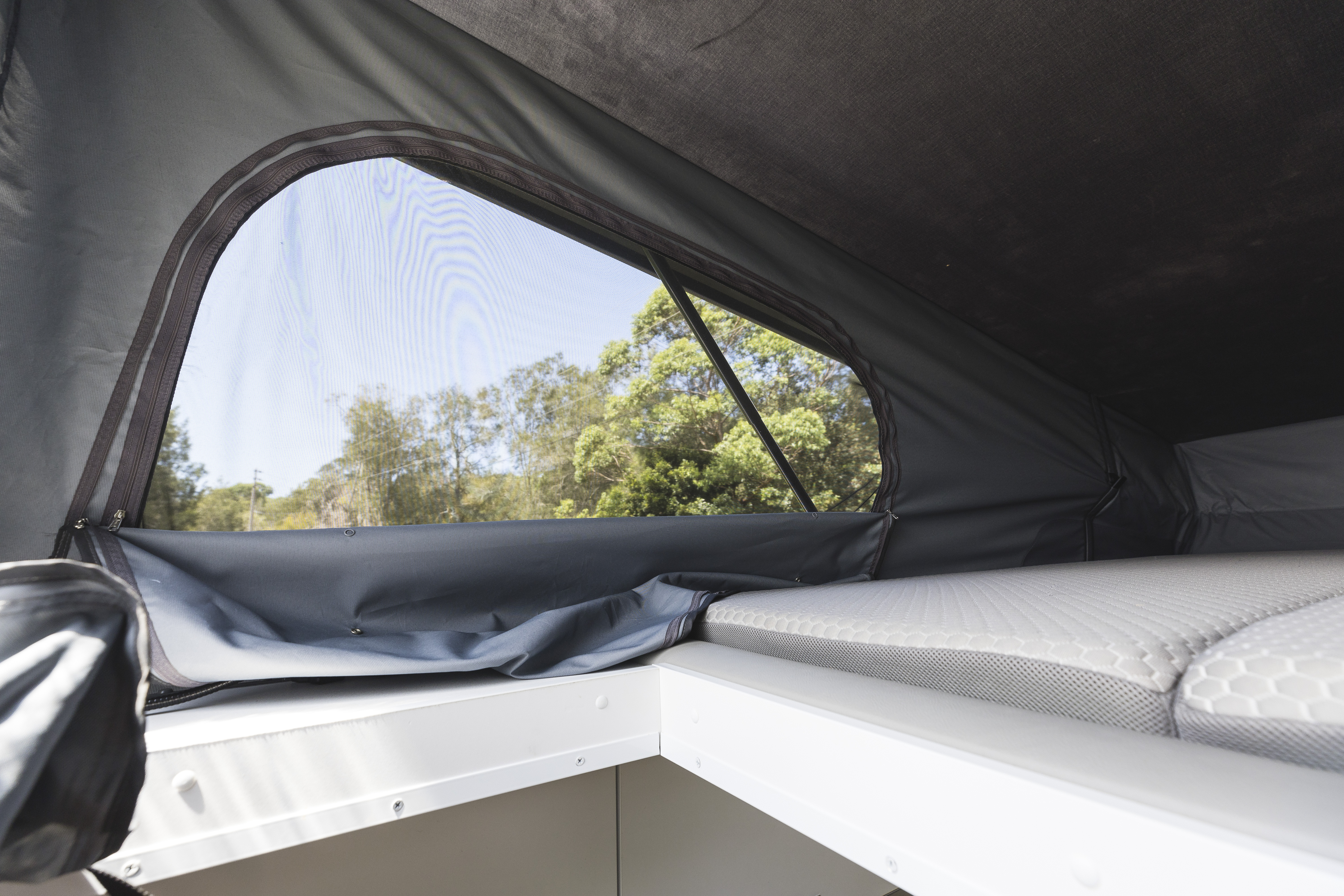 Interior view of a campervan roof tent with mattress and mesh window overlooking trees outside.